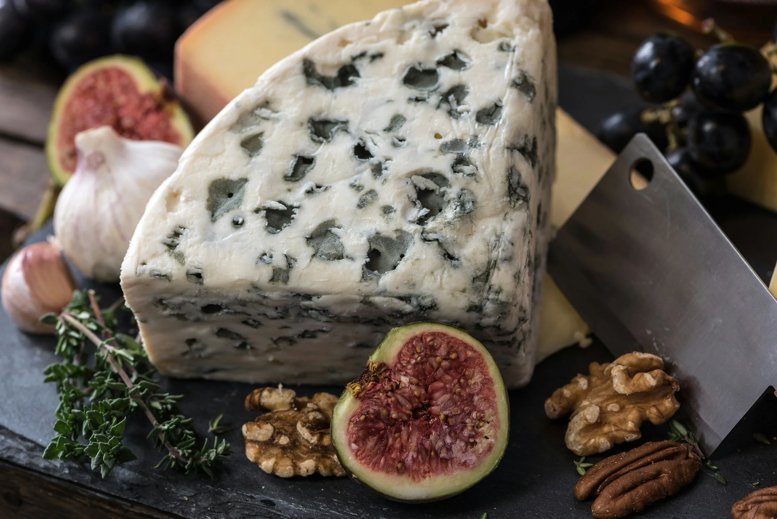 A wedge of blue-veined cheese surrounded by fresh figs, garlic, walnuts, and herbs on a dark slate serving platter.