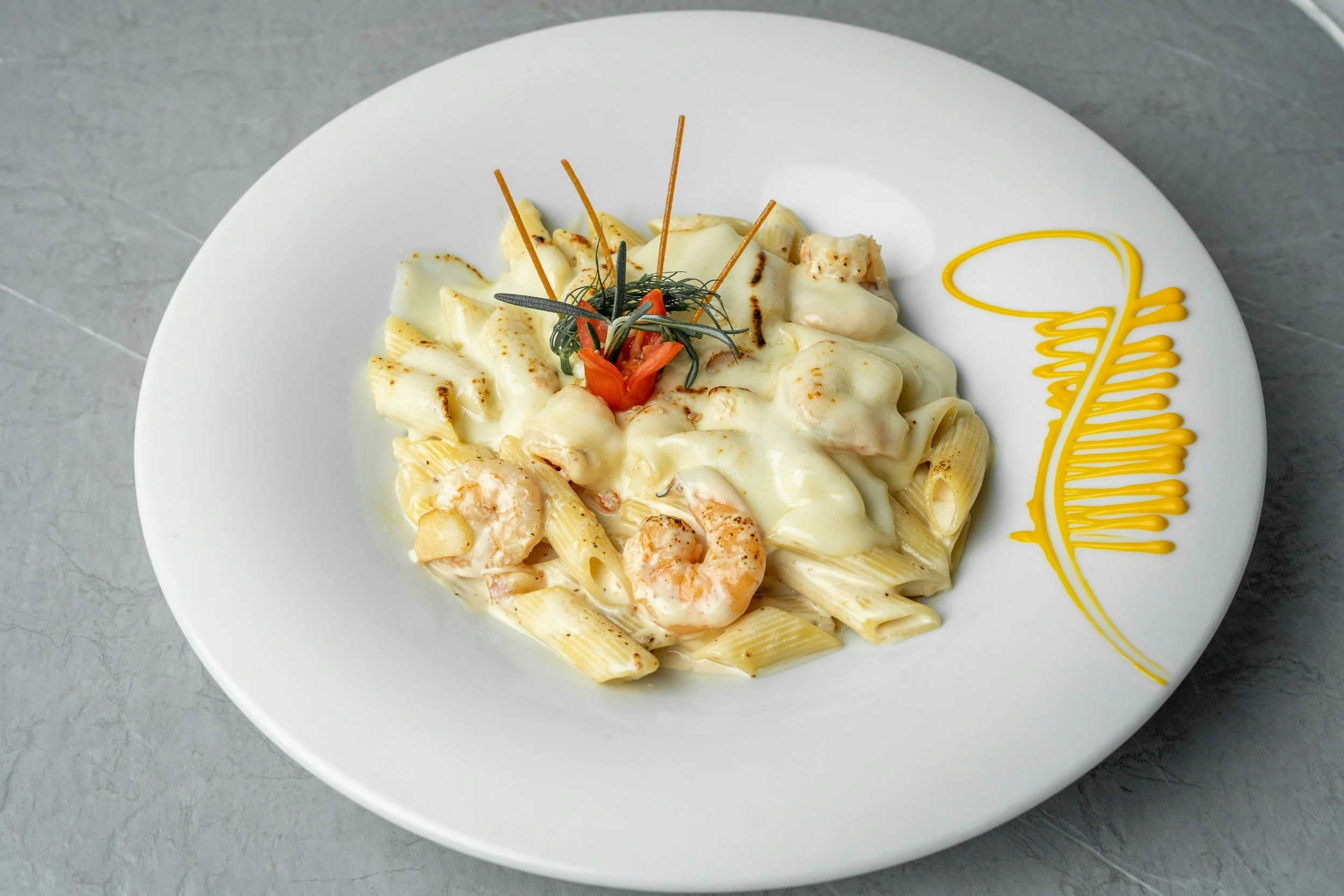 Plate of penne pasta with creamy white sauce and shrimp, garnished with herbs and peppers, served on a white dish with a yellow decorative sauce on the side.