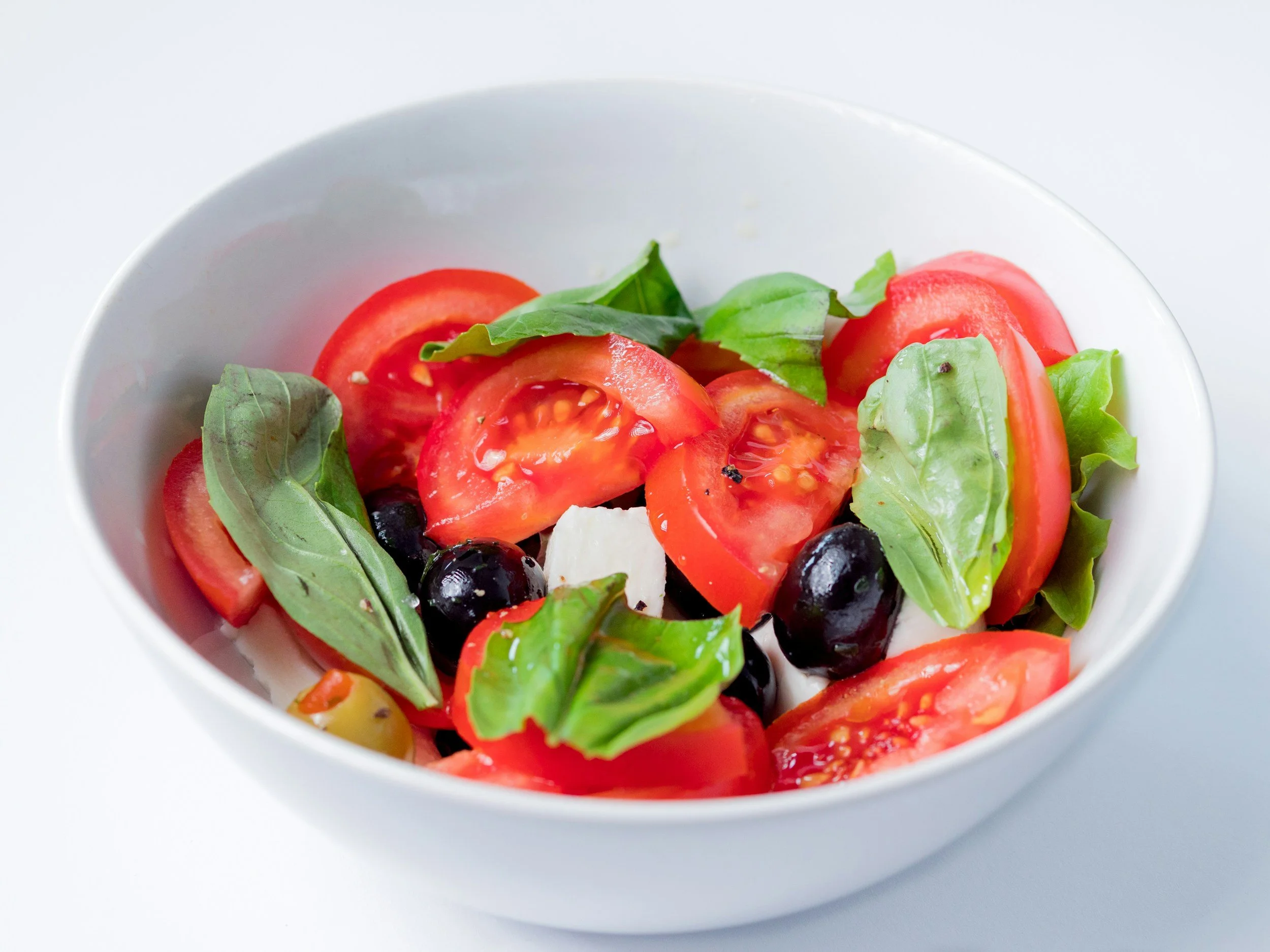 Bowl of fresh salad with sliced tomatoes, basil leaves, black olives, and mozzarella cheese.