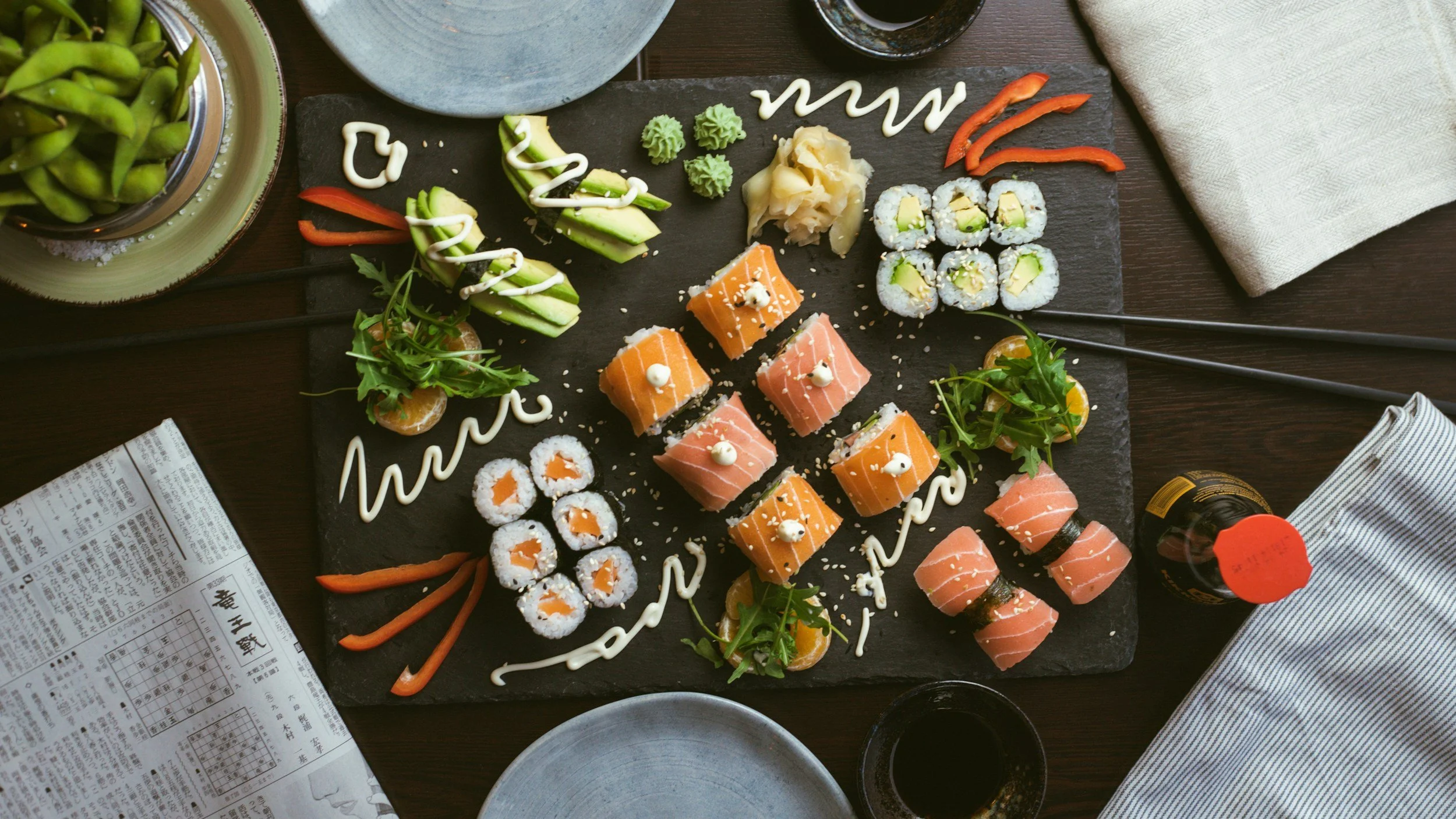 Assorted sushi rolls on a black stone platter with decorative sauces, garnishes, and wasabi, set on a dining table with plates, chopsticks, and a newspaper.