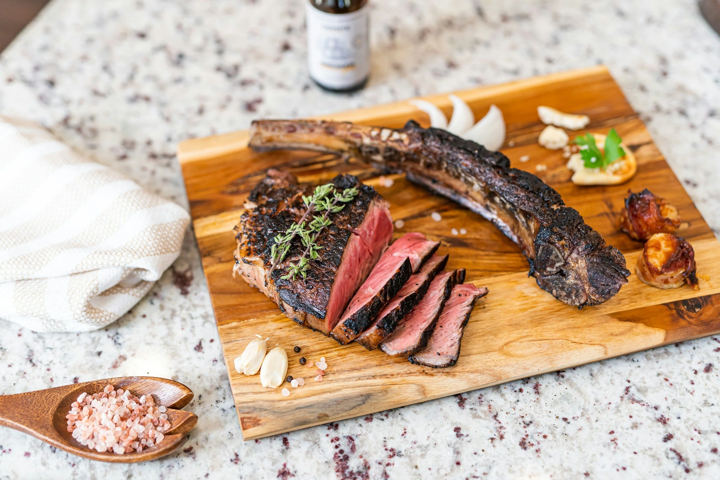A wooden cutting board with a cooked beef steak, a beef rib, grilled garlic, and seasoning salt on a granite countertop.