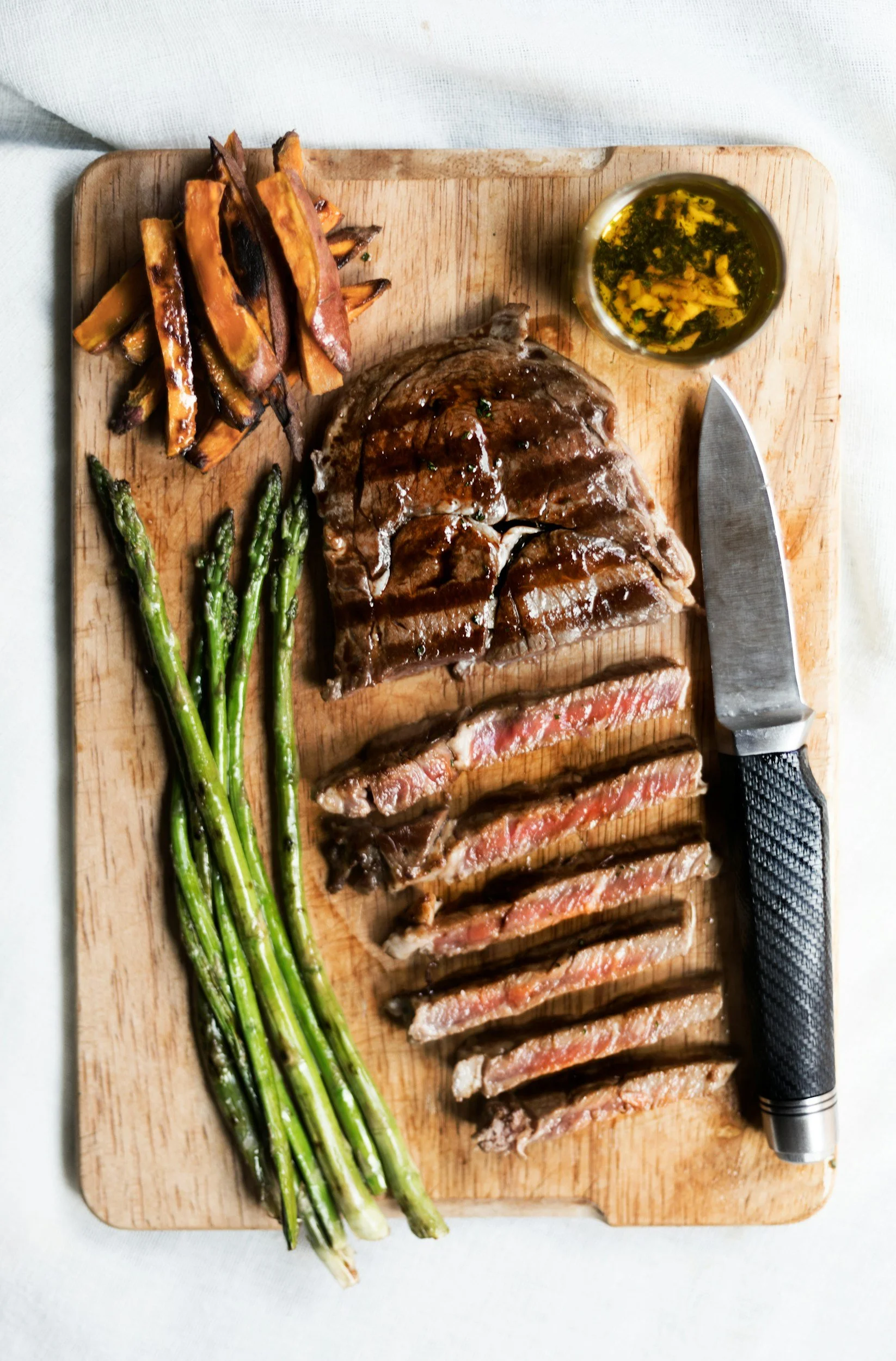 Grilled steak sliced on a wooden cutting board with roasted sweet potato fries, green asparagus, a small cup of sauce, and a side of dipping sauce.