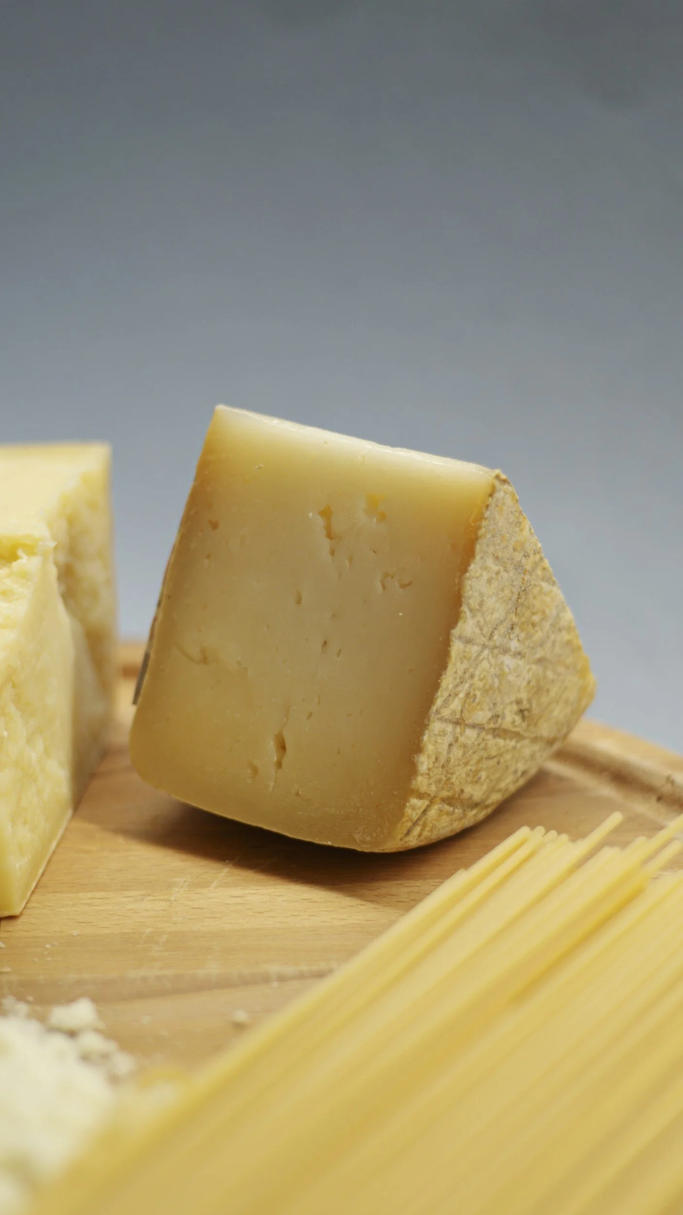Close-up of a wedge of hard yellow cheese on a wooden surface, with other cheese pieces and pasta in the background.