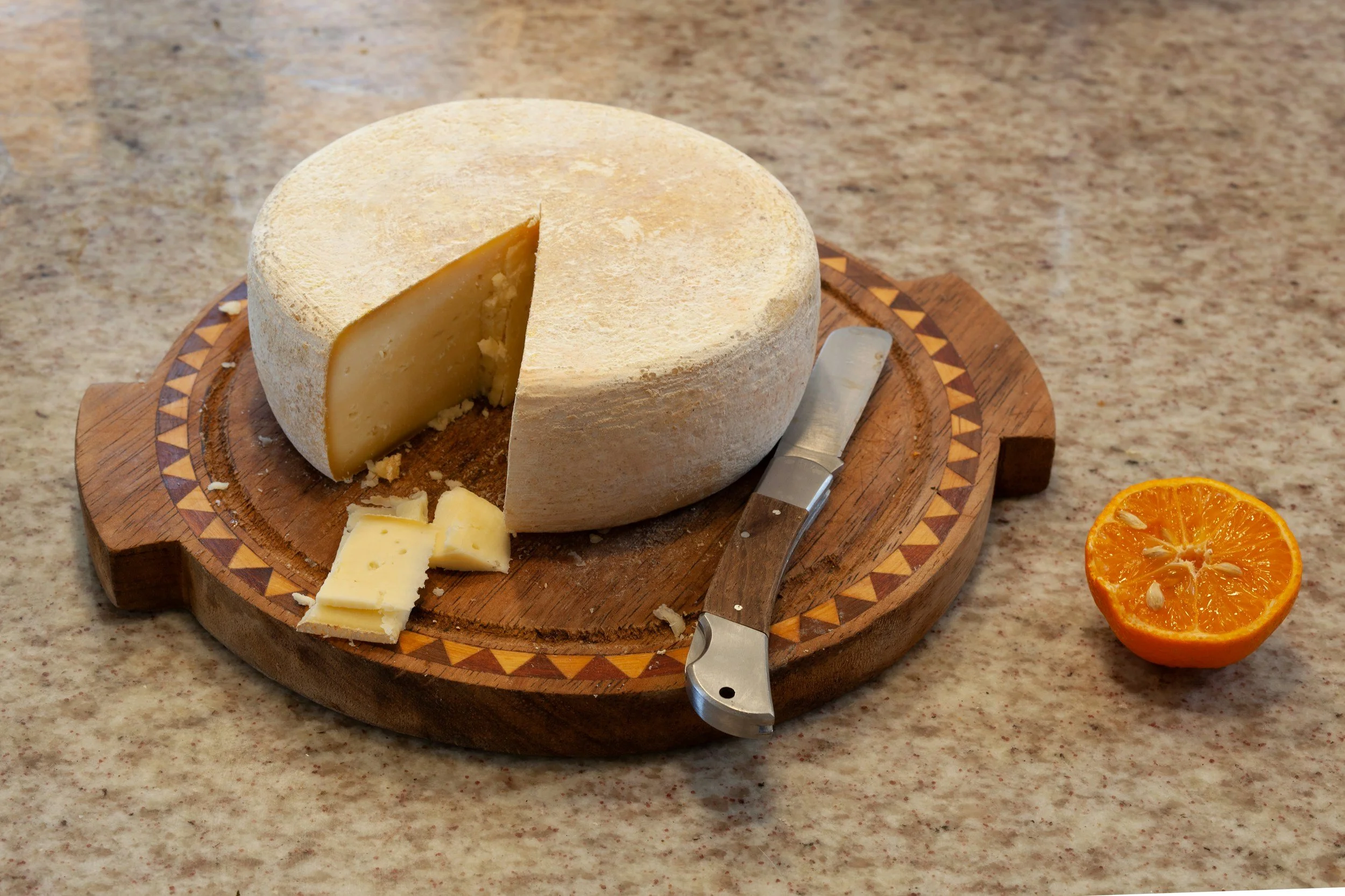 A round wheel of cheese with a slice cut out, placed on a wooden cutting board, next to a cheese knife, on a countertop. An orange halves sits to the right.