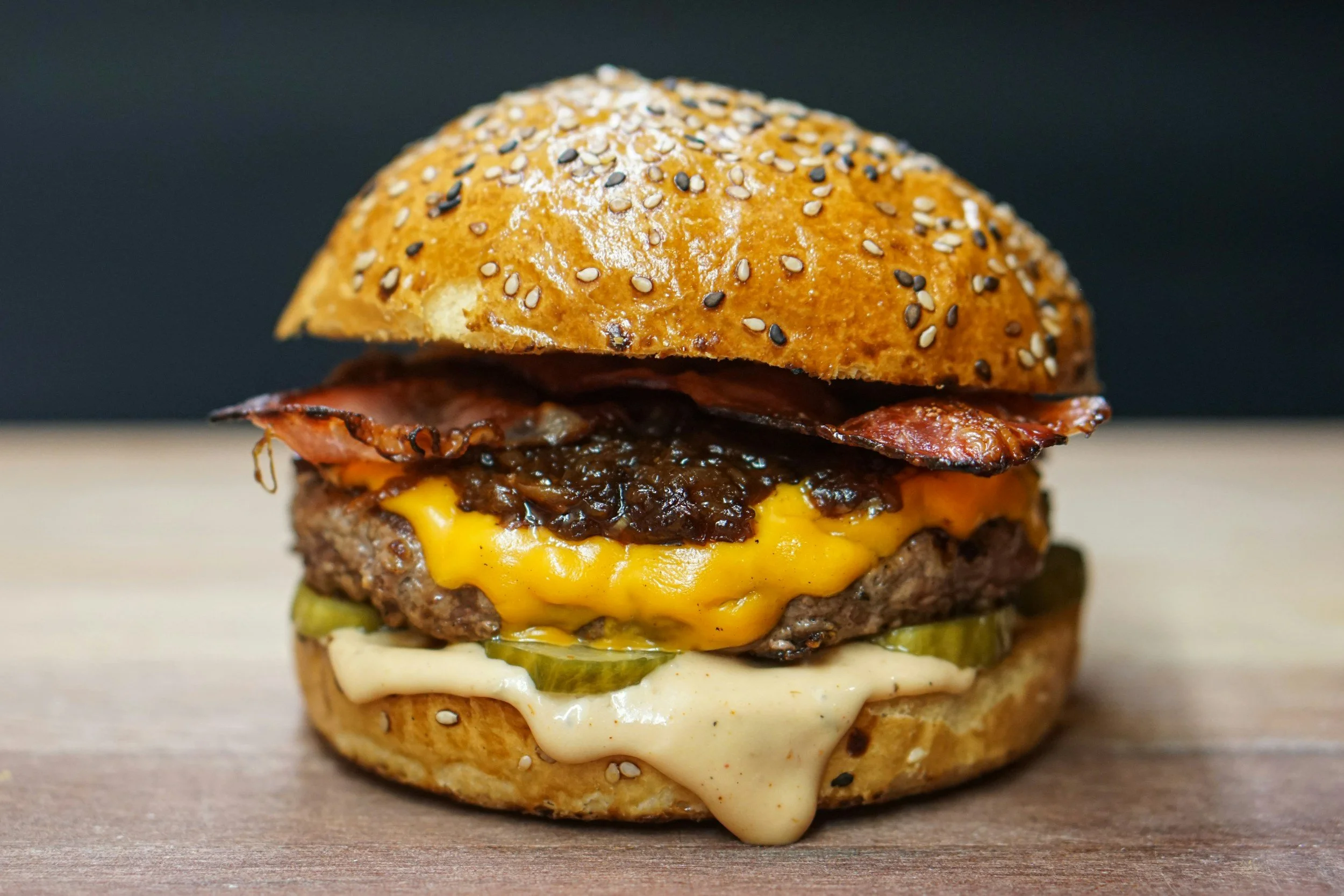 Close-up of a cheeseburger with sesame seed bun, cheddar cheese, bacon, pickles, and sauce.