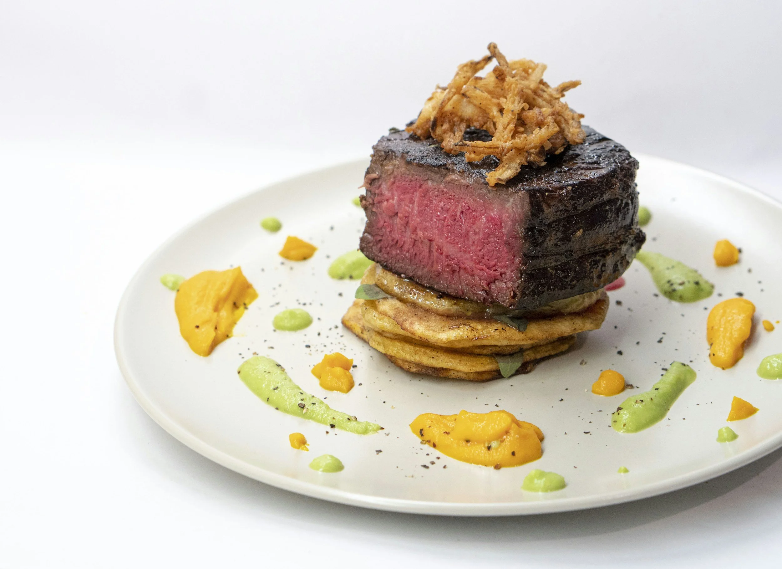 A plated dish featuring a medium-rare steak topped with crispy onion strings, served on a bed of roasted vegetables with colorful sauce dollops and garnished with black pepper.