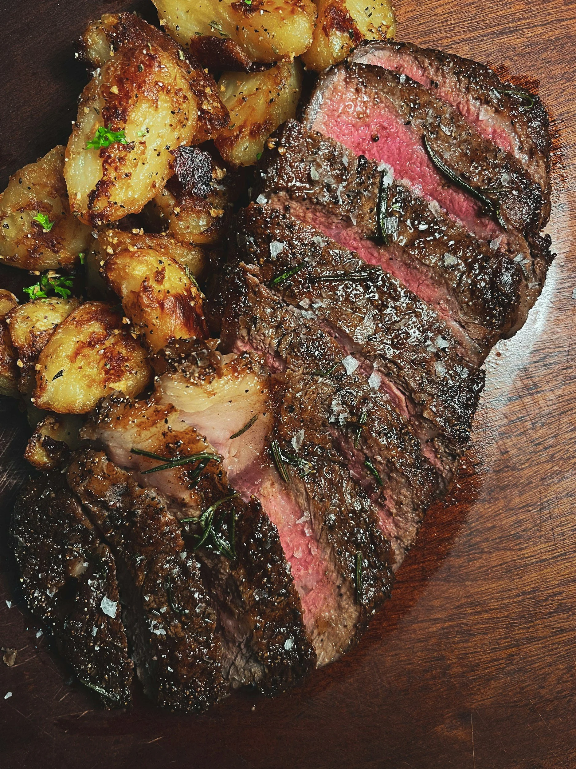 Slices of cooked steak with a pink center and roasted potatoes garnished with herbs on a wooden cutting board.