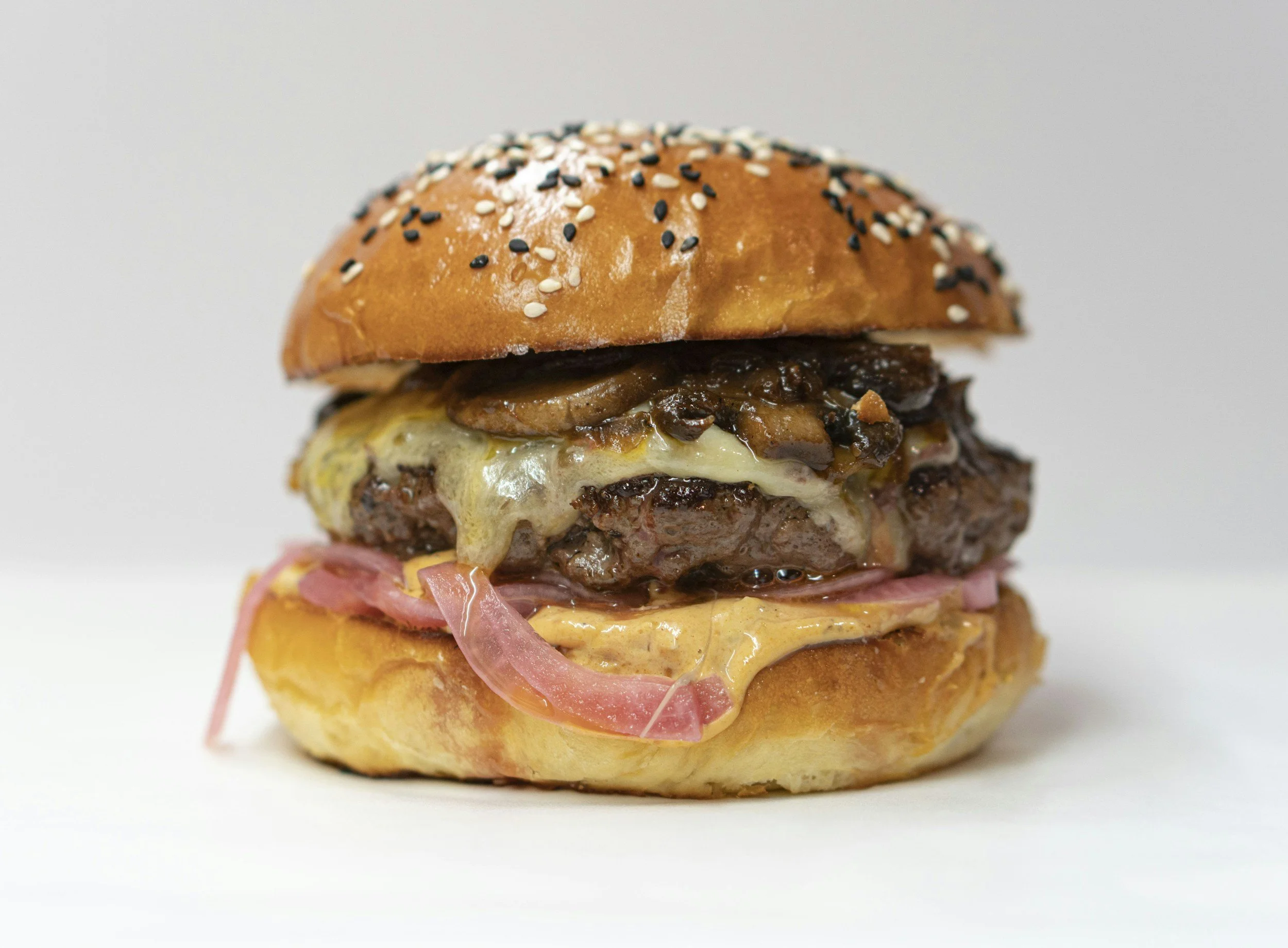 Close-up of a cheeseburger with a sesame seed bun, cheddar cheese, grilled onions, sliced ham, and a beef patty on a white background.