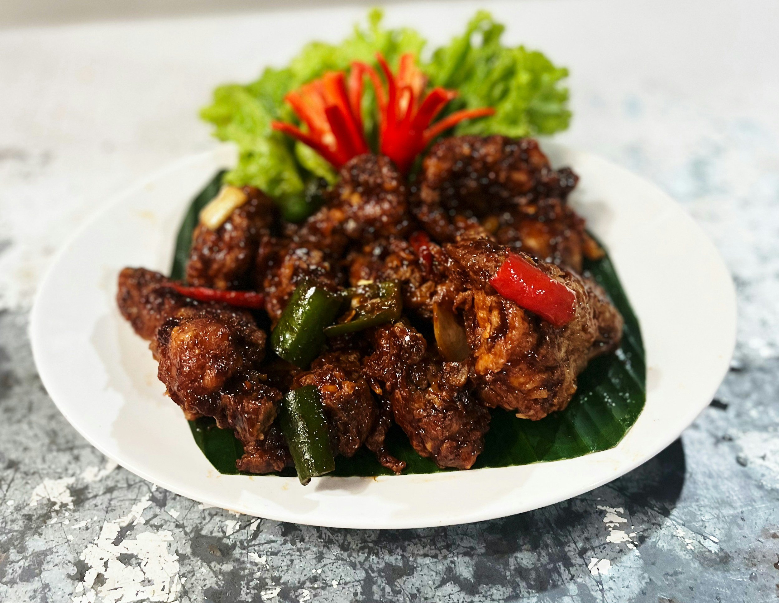 Plate of cooked spicy chicken with red and green peppers, garnished with lettuce and red bell pepper slices, on a white dish with green leaf underneath, on a gray textured surface.