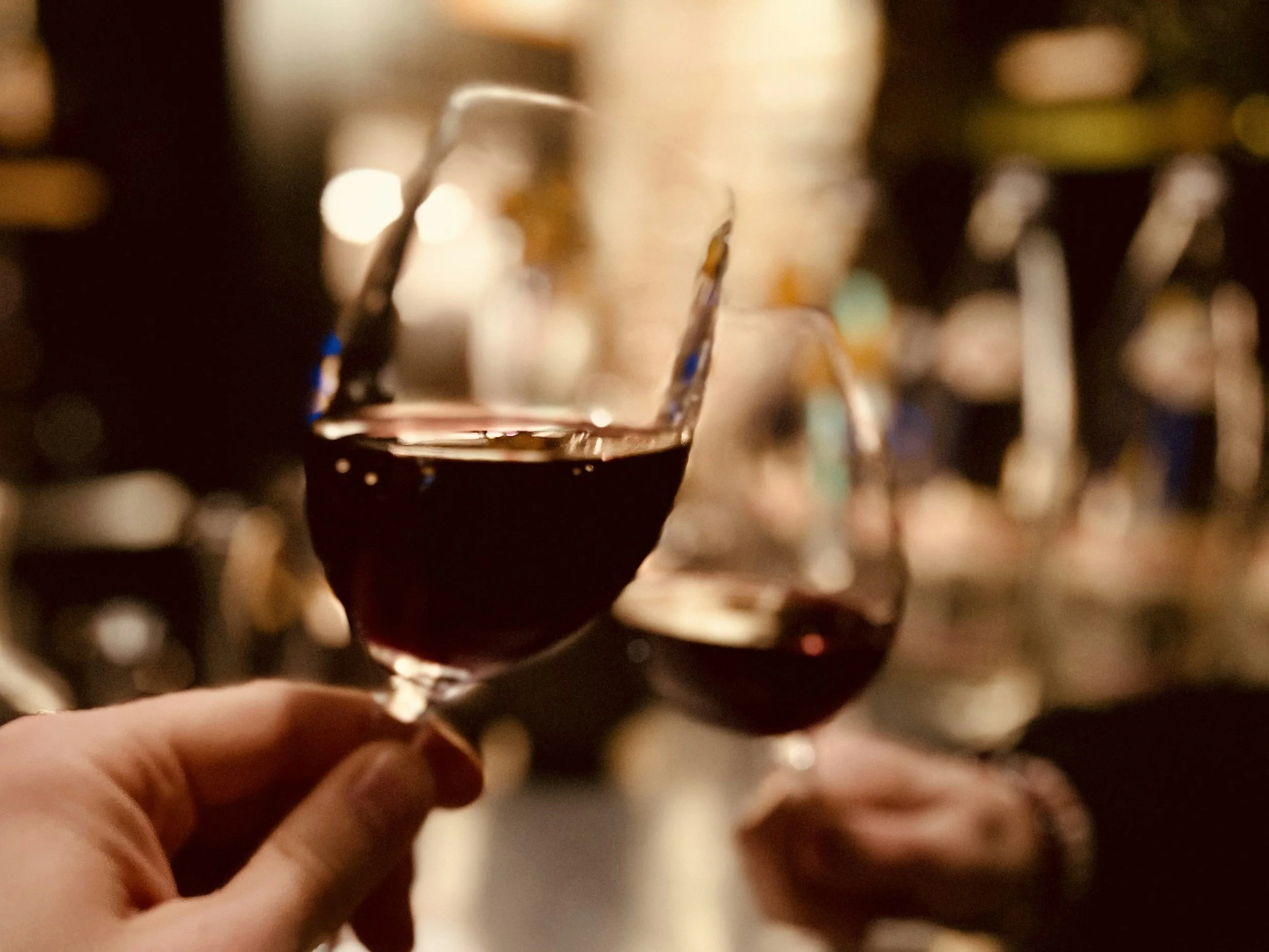 Two glasses of red wine being held in front of a bar with blurred bottles in the background.