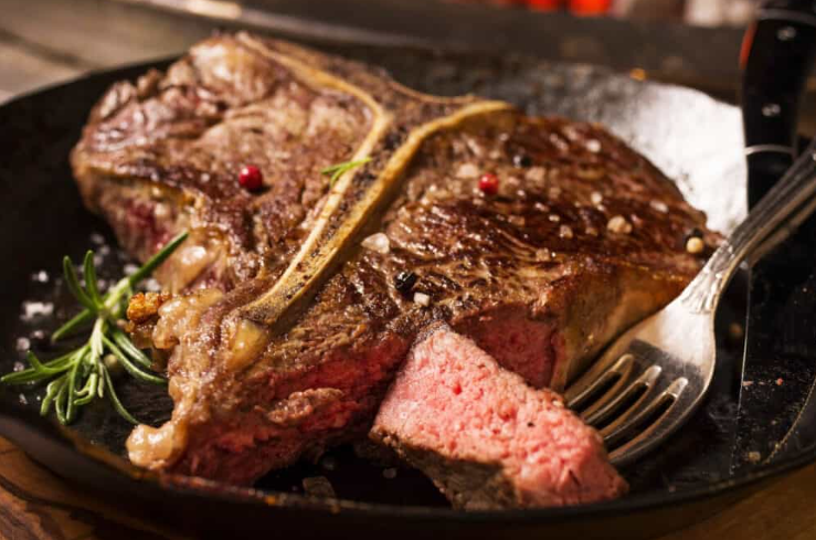 A cooked prime rib steak on a black skillet with a sprig of rosemary, pink interior meat, and a silver fork on the side.