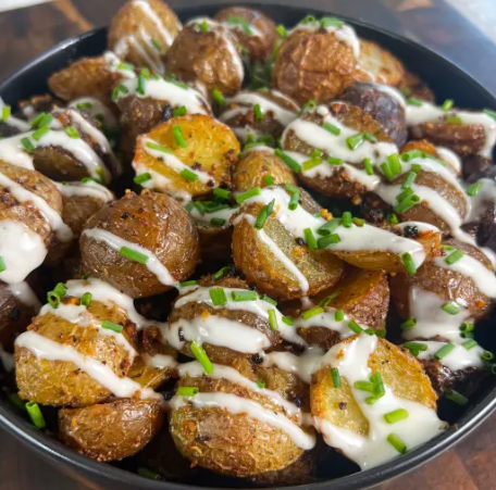 A black bowl filled with roasted baby potatoes topped with sour cream, chopped chives, and drizzled with a sauce.