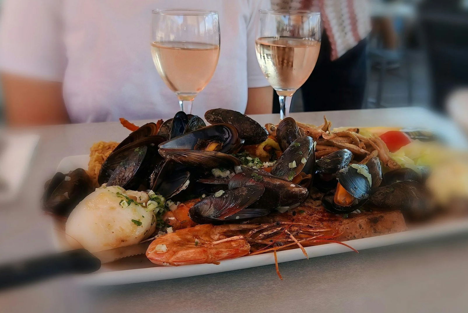 Seafood platter with mussels, prawns, fish, and side dishes, served on a white rectangular plate, accompanied by two glasses of white wine, on a white table.