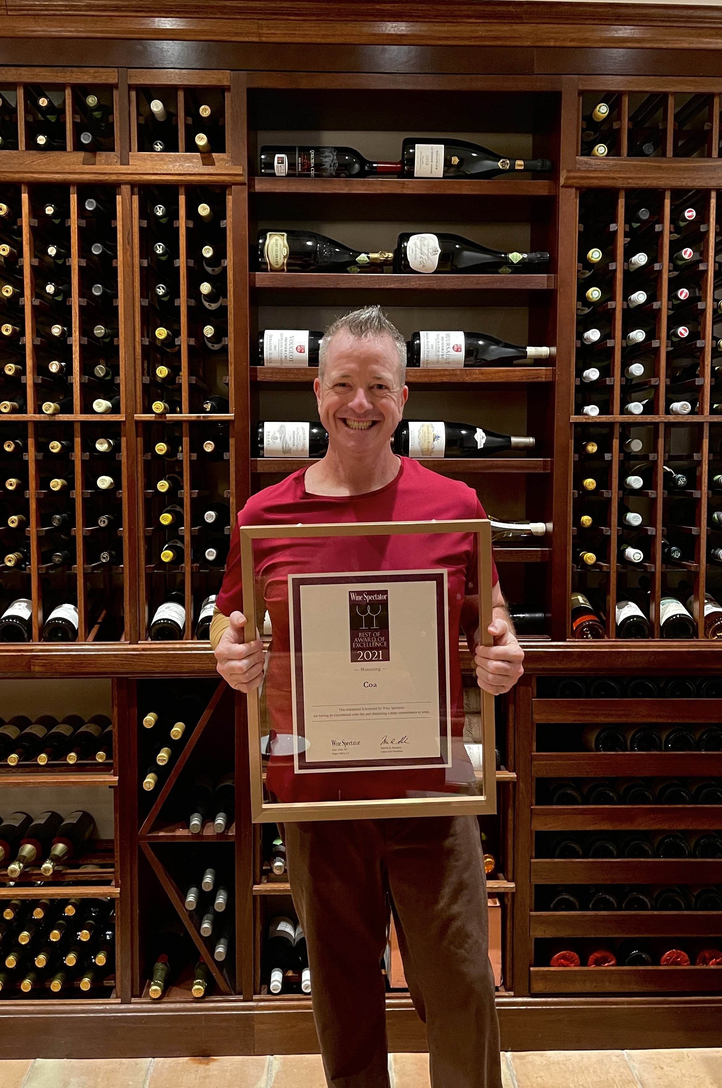 Man holding award certificate in front of a large wine rack filled with various bottles.