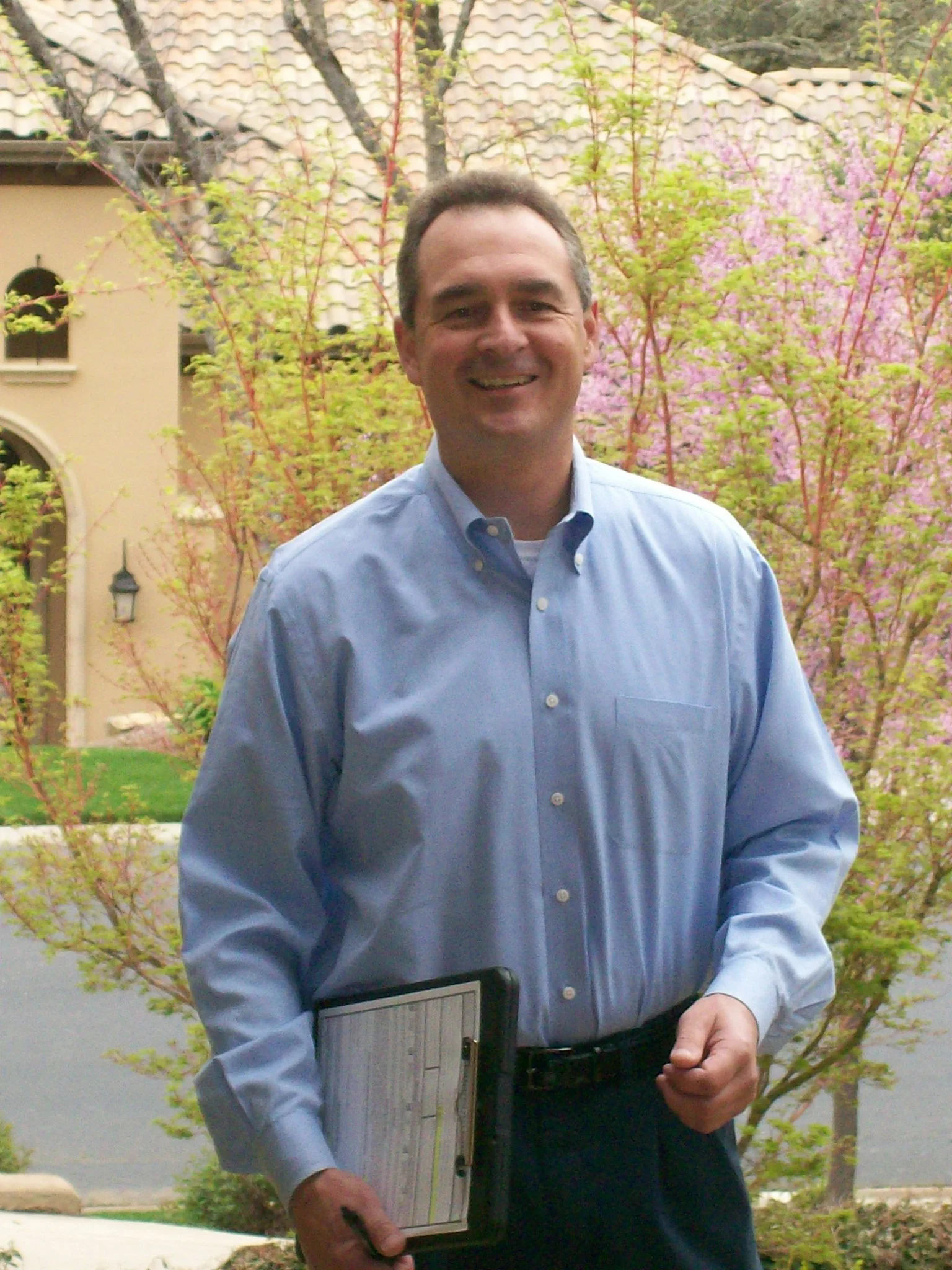 A man in a blue dress shirt holding a clipboard, standing outdoors with pink flowering trees and a house with a tiled roof in the background.