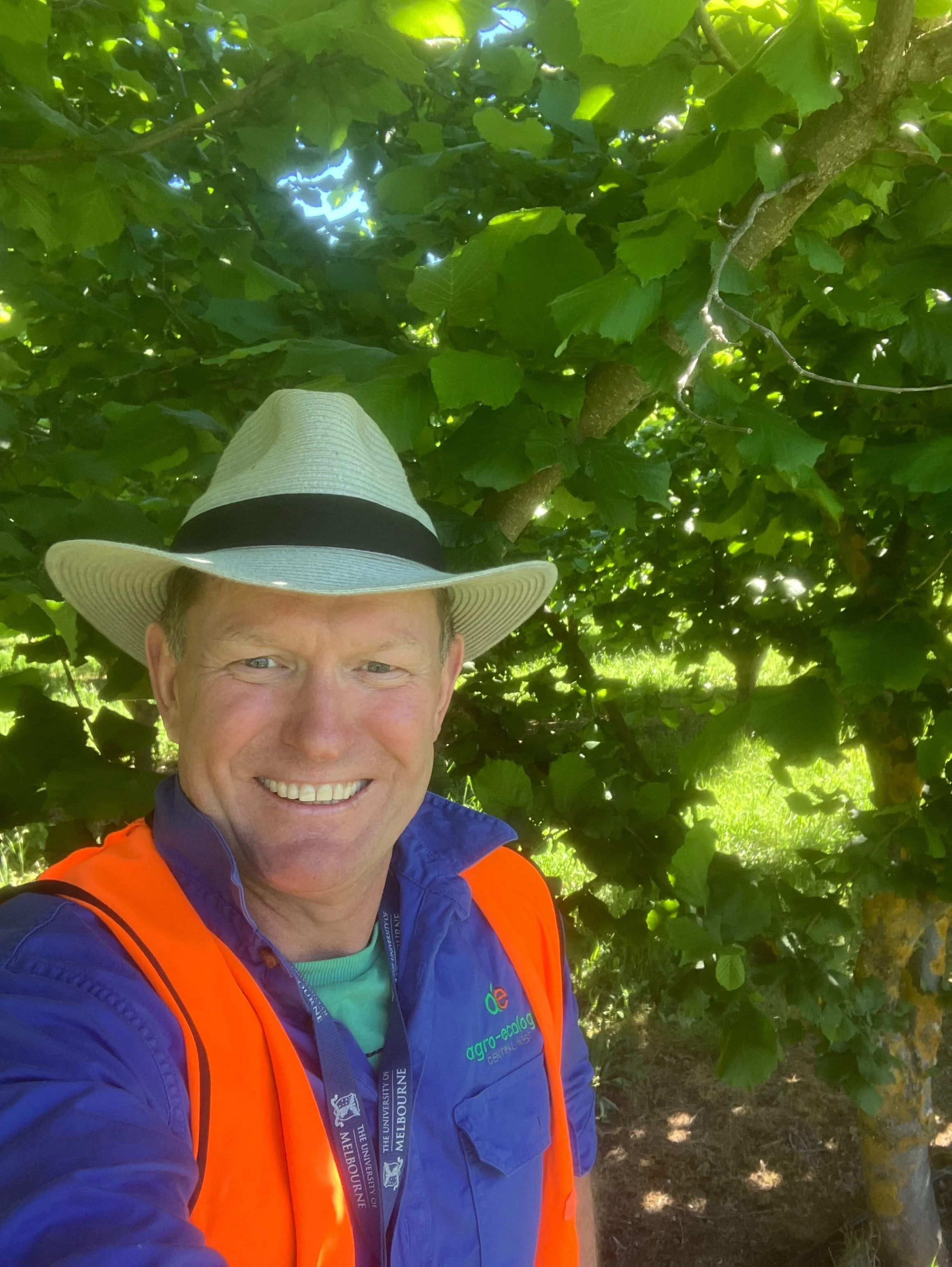 A man wearing a wide-brimmed hat, a blue jacket with a logo, and an orange safety vest, smiling for a selfie in a lush green outdoor setting.