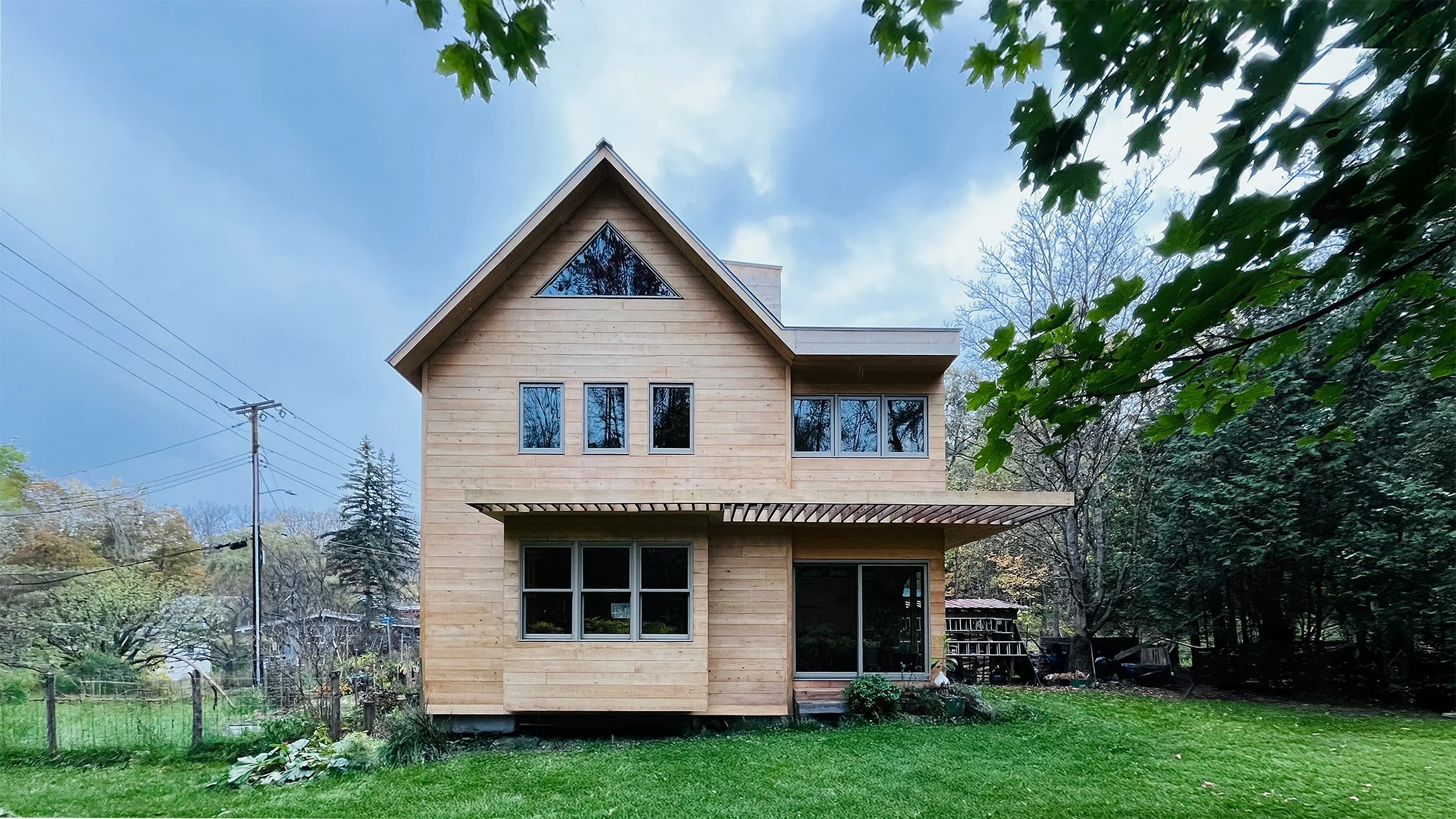 A warm, contemporary cedar clad home addition and renovation with a modern pergola trellis, expanded bay window, and refined window composition — a bespoke project that advances the archetypal gable box forms of New England villages, designed by Brya