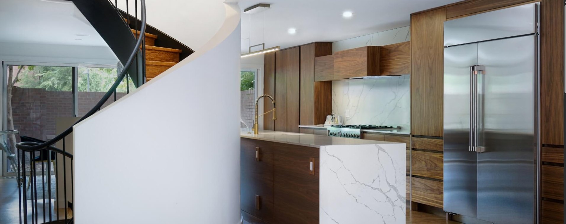 Interior view of a modern walnut kitchen with a marble island and a black metal spiral staircase curving upward beside it