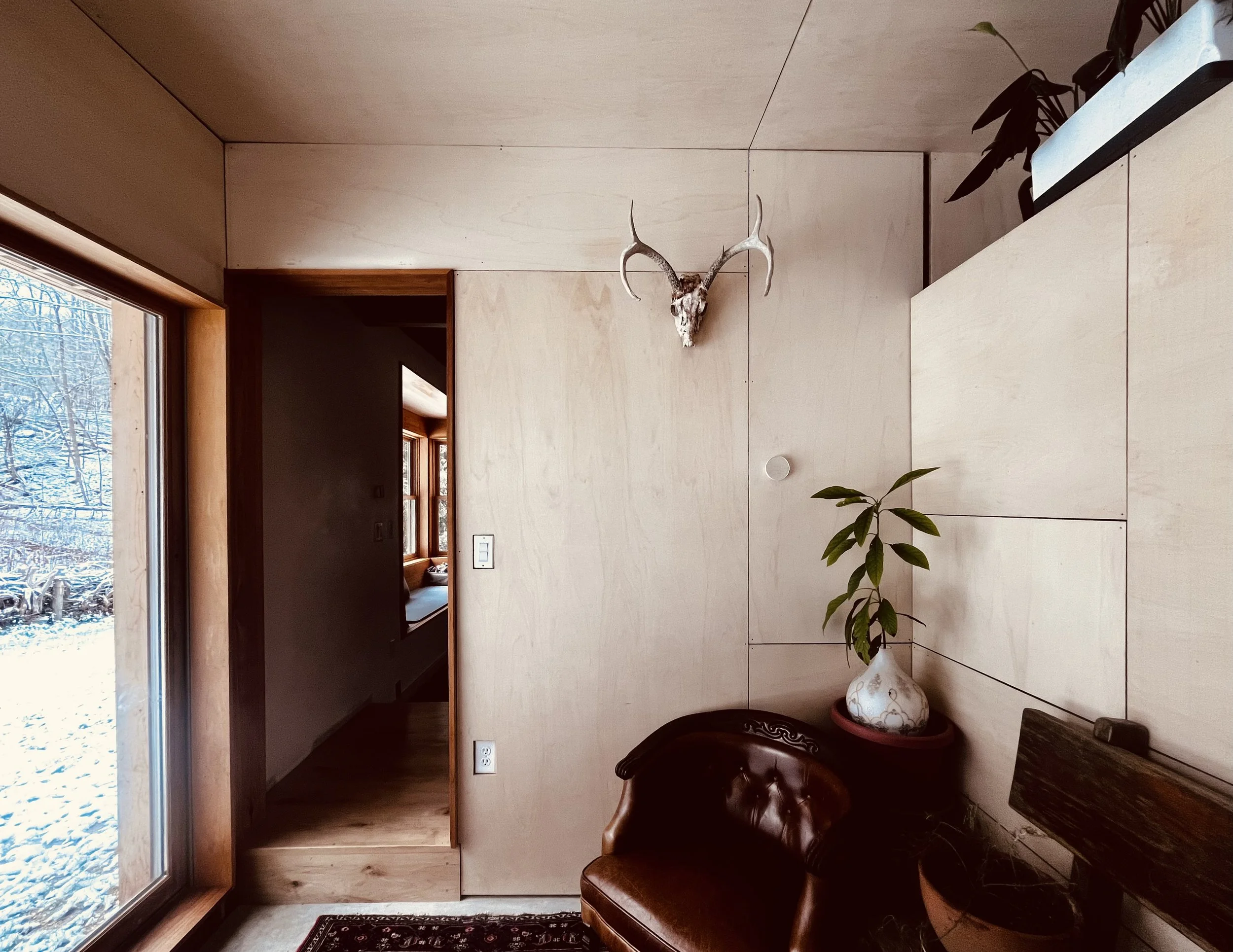 Sunroom interior with plywood wall and ceiling paneling, a leather armchair, potted plants, and a snowy landscape visible through a large window.