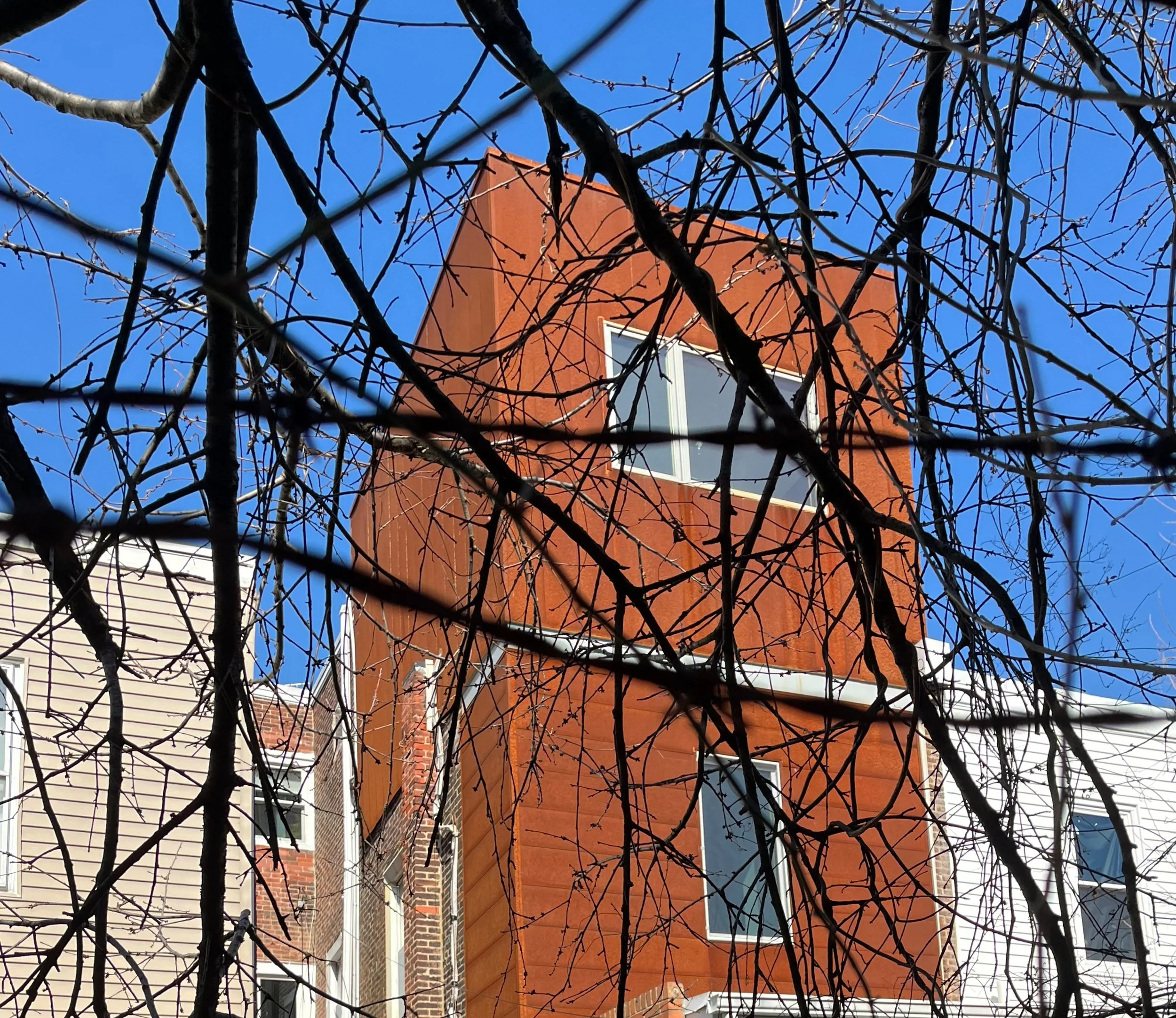 Compact corten steel rooftop addition with a sharply angular roofline, seen through leafless branches against a blue sky.