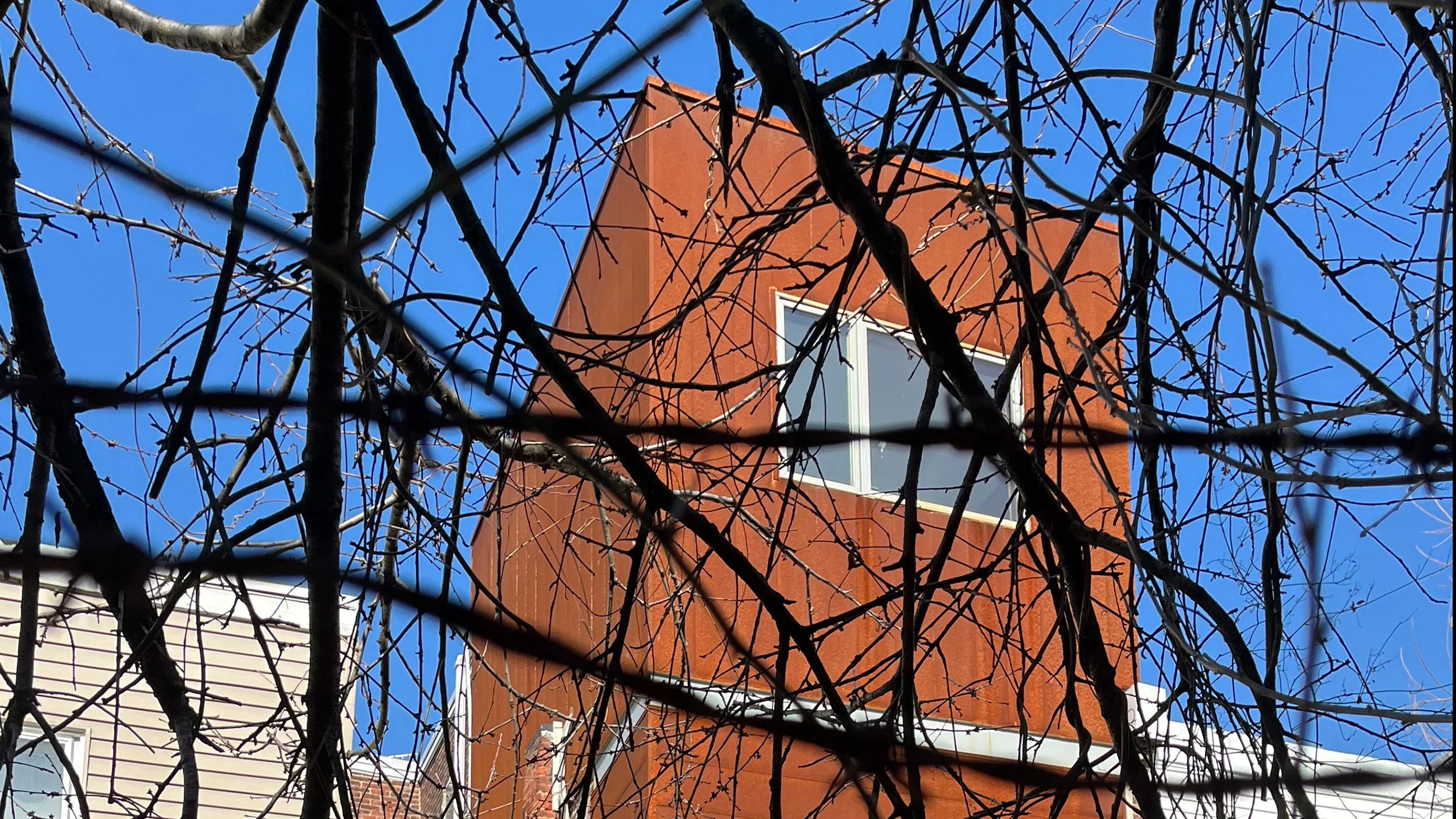 A compact, cubic corten-steel addition rising boldly from the rear of a Philadelphia rowhouse, featuring a sharply angular roofline and crisp contemporary detailing that at once contrasts with and befits the home as a carbon-colored cousin to its his