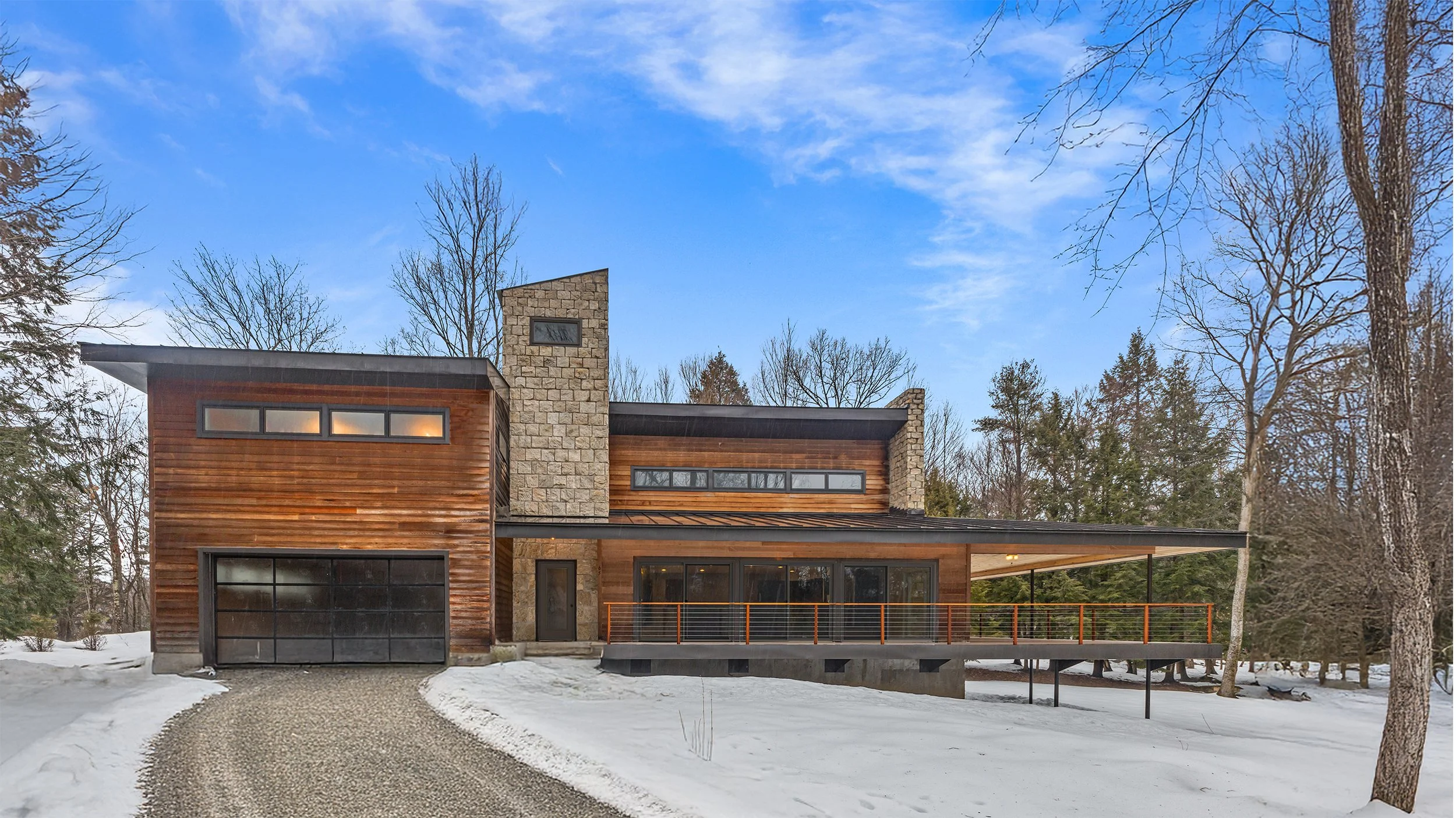 A sculptural, contemporary Vermont woodland residence featuring a stone tower, expansive cantilevered porch with an outdoor fireplace, horizontal cedar siding, and cedar cable railing. Designed by architect Bryan D. Jones of Skyplane Studio Architect