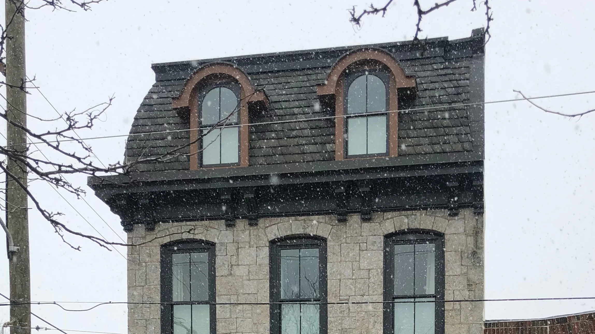 A historic Second Empire–style three story stone building with a mansard roof, arched dormer windows, and fish scale slate tiles stands at a snowy West Philadelphia street corner.