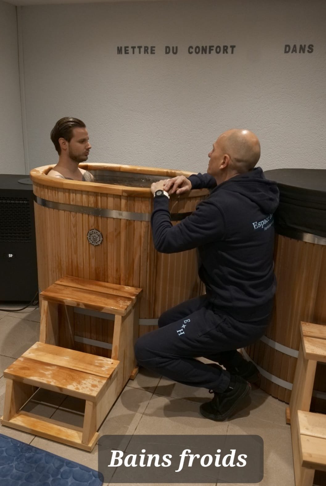 Un homme sans vêtements dans un petit spa gonflable noir, en plein air, devant une vieille maison en pierre avec volets bleus. Le soleil brille et la nature verdoyante entoure la scène.