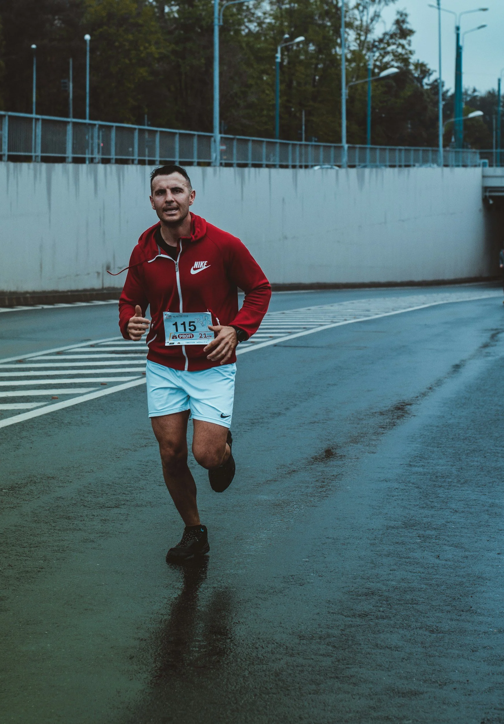 Un homme court lors d'une course en extérieur, portant un maillot rouge Nike, un short blanc, et une ceinture avec un numéro de course 115 sur son ventre, sous un ciel gris, sur une route mouillée.