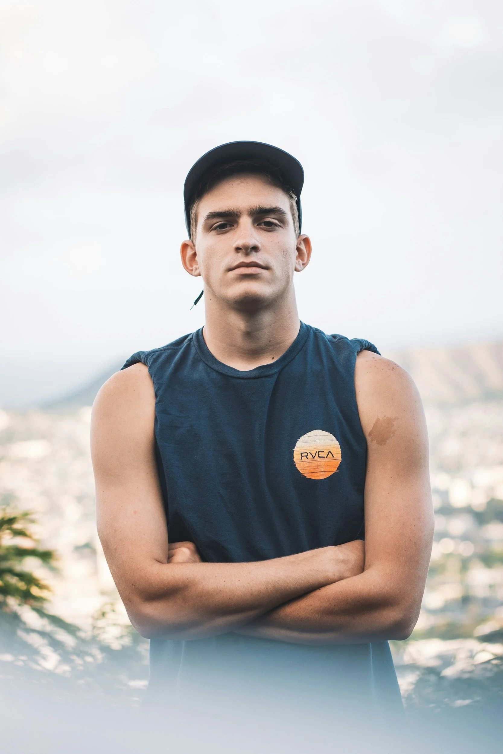 Jeune homme avec bras croisés, portant un t-shirt sans manches et une casquette, devant un paysage urbain lors d'une journée nuageuse.