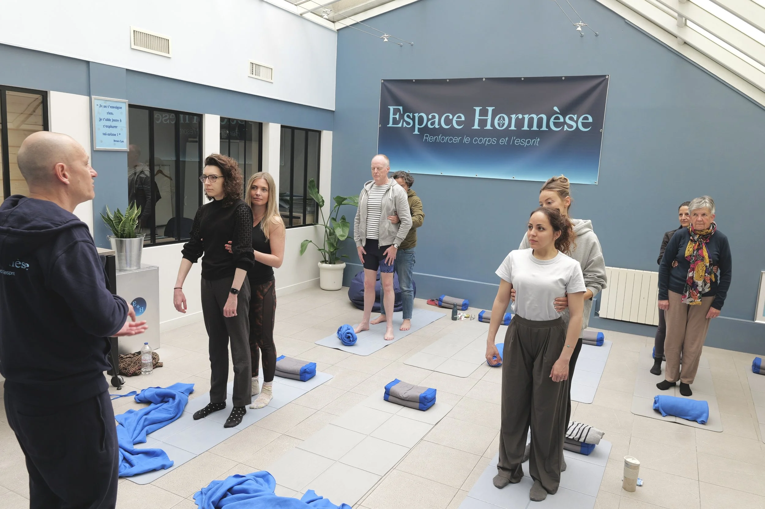 Groupe de personnes allongées sur un tapis de yoga dans une salle avec de grandes fenêtres, un instructeur en pied.