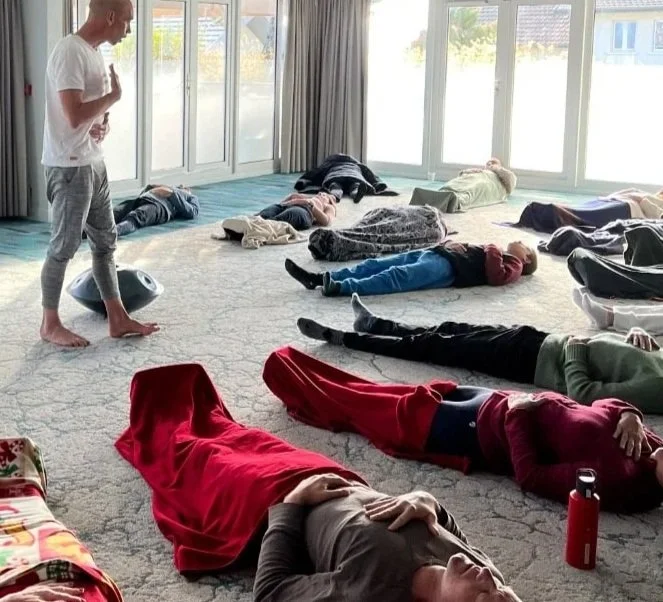 Groupe de personnes allongées sur le sol, participate à une séance de relaxation ou méditation dans une pièce lumineuse avec de grandes fenêtres.