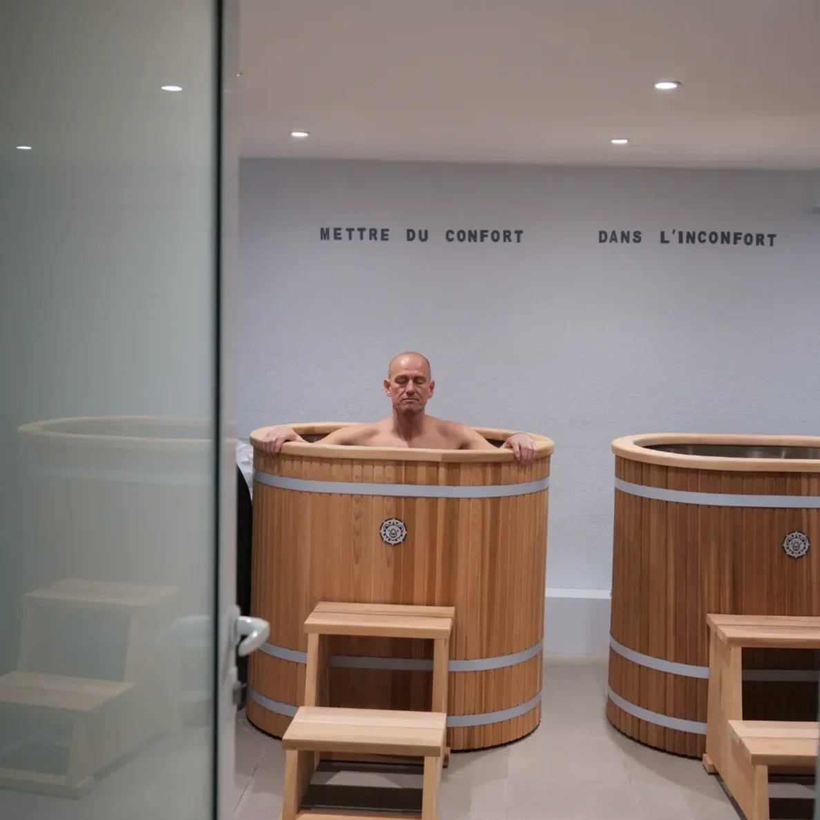 Un homme sans vêtements dans un petit spa gonflable noir, en plein air, devant une vieille maison en pierre avec volets bleus. Le soleil brille et la nature verdoyante entoure la scène.
