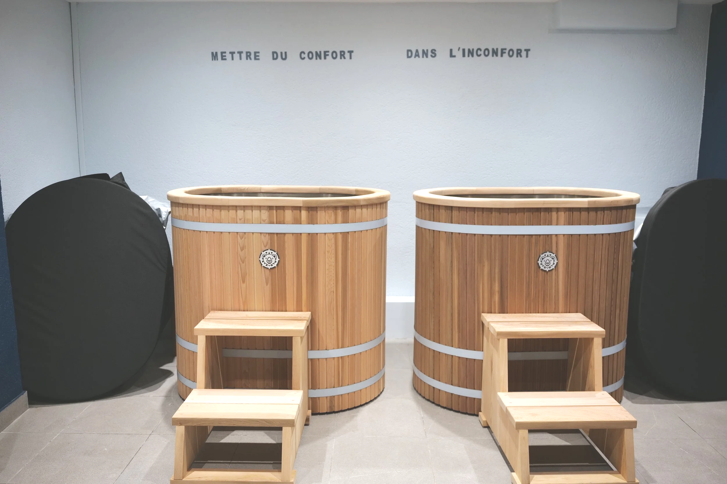 Deux baignoires en bois avec deux petites escaliers en bois devant, sur un fond de mur blanc avec l'inscription "Mettre du confort dans l'inconfort" en lettres noires.