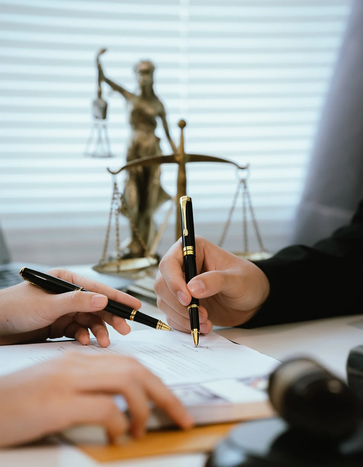Two people signing documents at a desk with a statue of Lady Justice in the background.