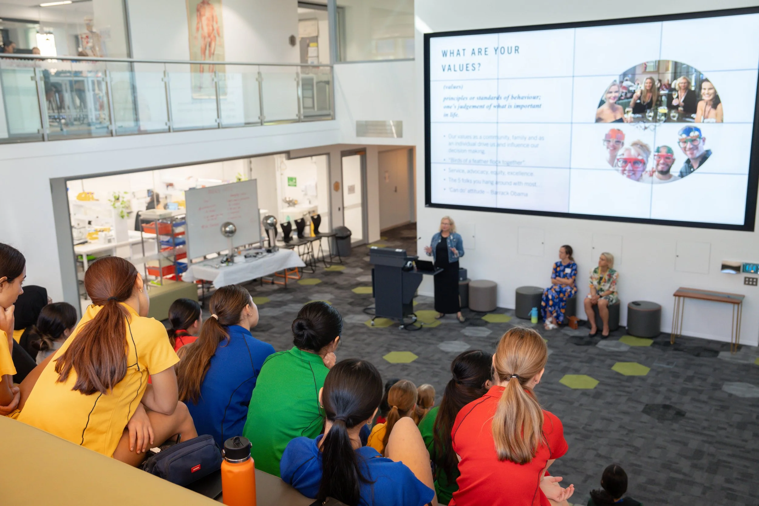 A woman giving a presentation to a group of women and girls seated in a modern, multi-level conference room. The presentation slide on a large screen reads 'What are your values?' with images of smiling people.