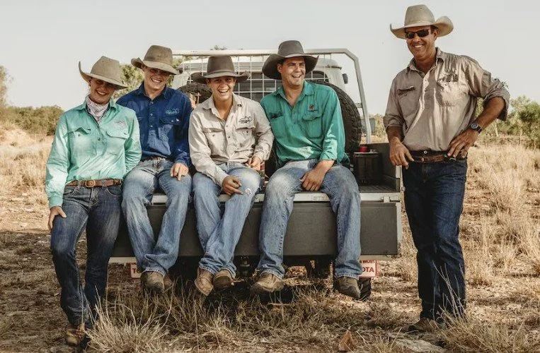 Five people dressed in cowboy hats and casual outdoor clothes posing next to a pickup truck in a dry field.