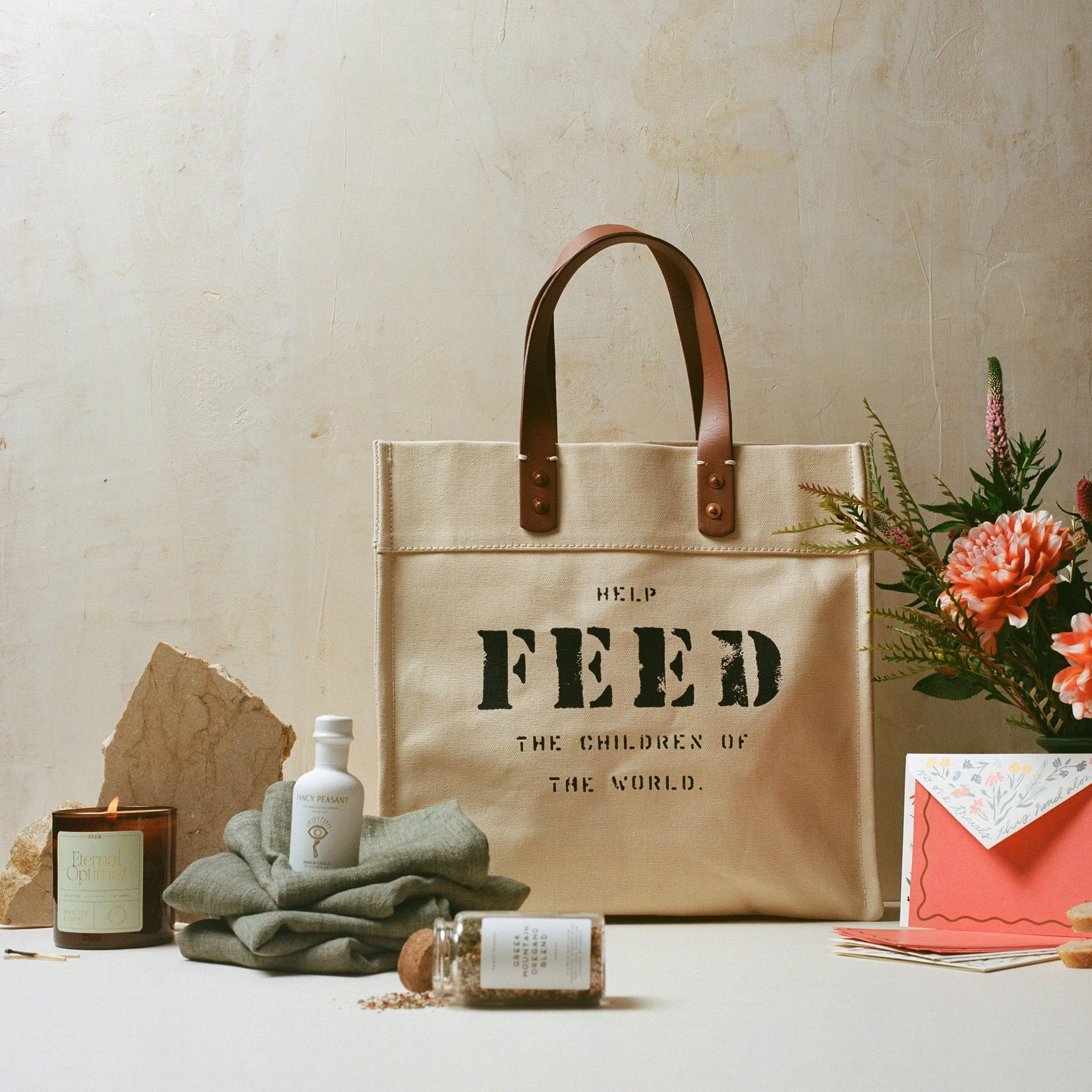 A beige tote bag with brown leather handles and black text reading 'Help Feed the Children of the World' on a table. Surrounding the bag are a candle, a small bottle, a folded gray cloth, a pink card, a flower arrangement, and a bottle of bath salts,