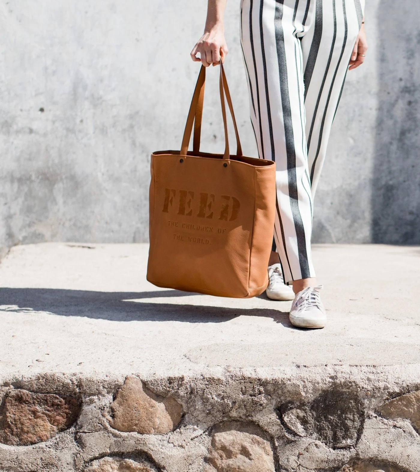 Person wearing black and white striped pants holding a brown tote bag with the word "FEED" printed on it, standing on a stone surface with a concrete wall in the background.