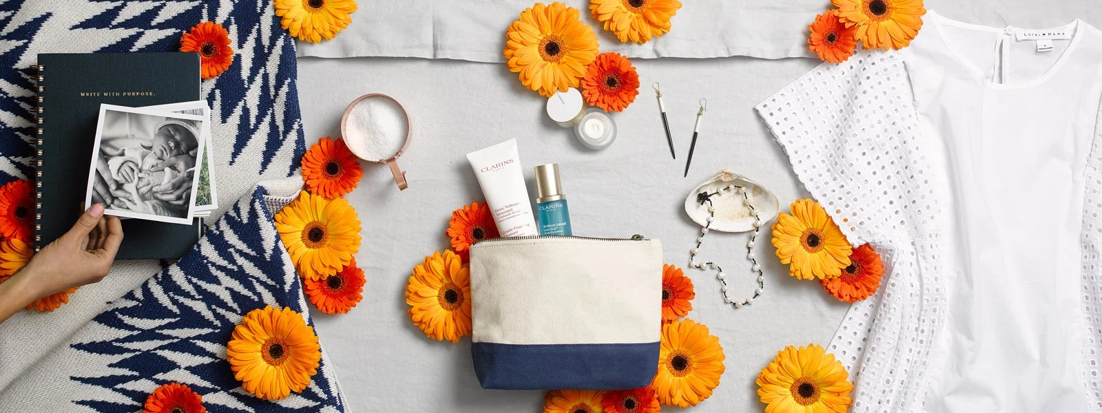 Flat lay of personal items on a white surface, surrounded by orange and yellow flowers, including a striped blue and white blanket, a white dress, skincare products, jewelry, a pouch, and black earrings.