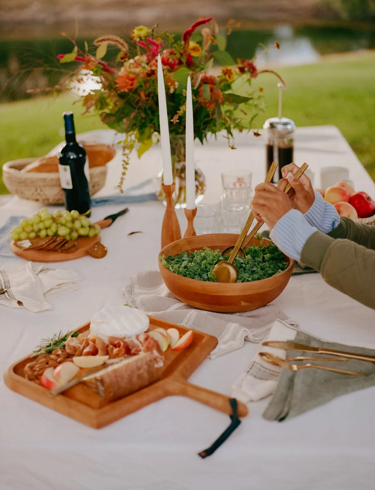 Outdoor dining table with a large flower arrangement, wine bottle, grapes, bread, salad, cheese, apples, and candles.