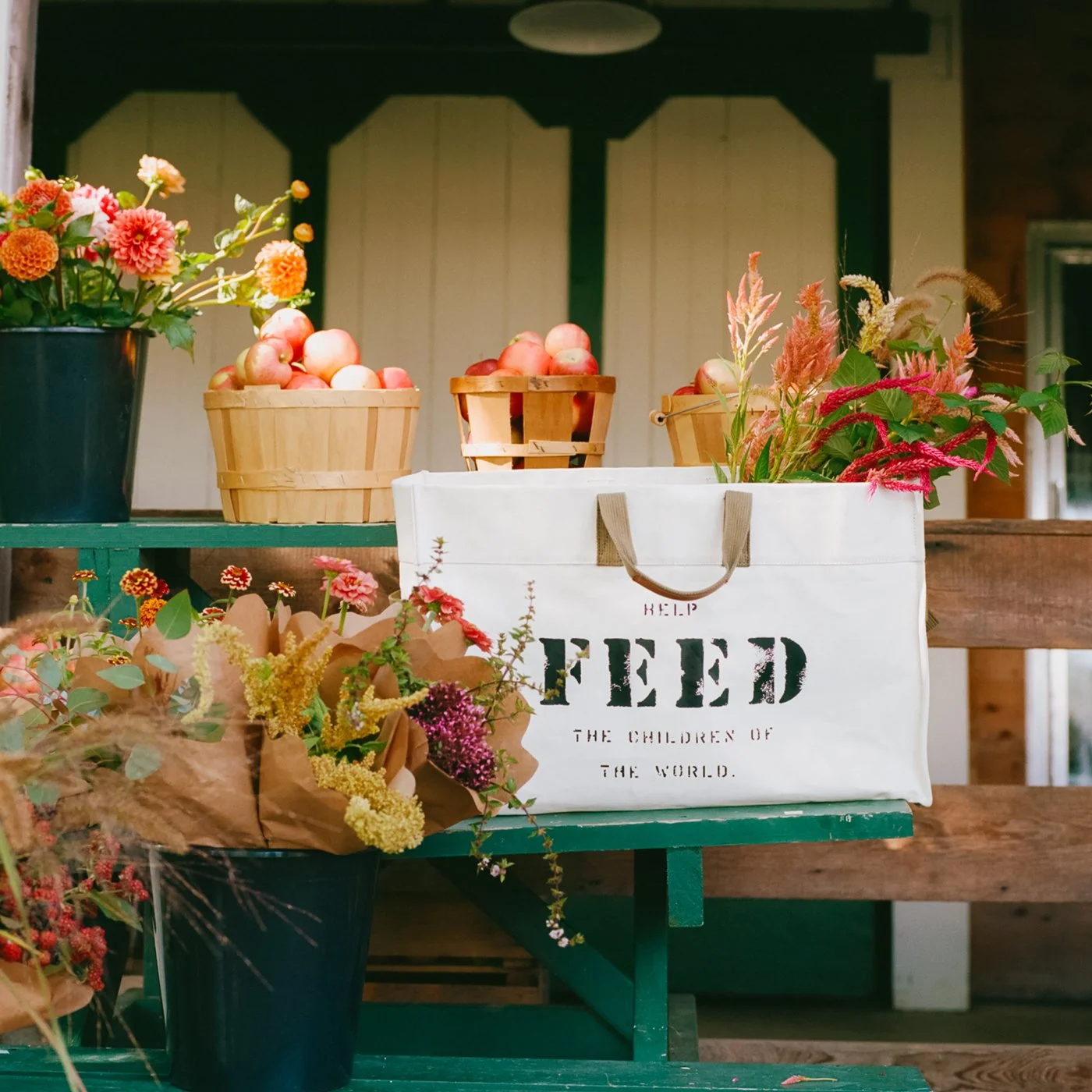 A flower and produce stand with baskets of apples, bouquets of flowers, and a white reusable bag with black text reading 'HELP FEED THE CHILDREN OF THE WORLD.'