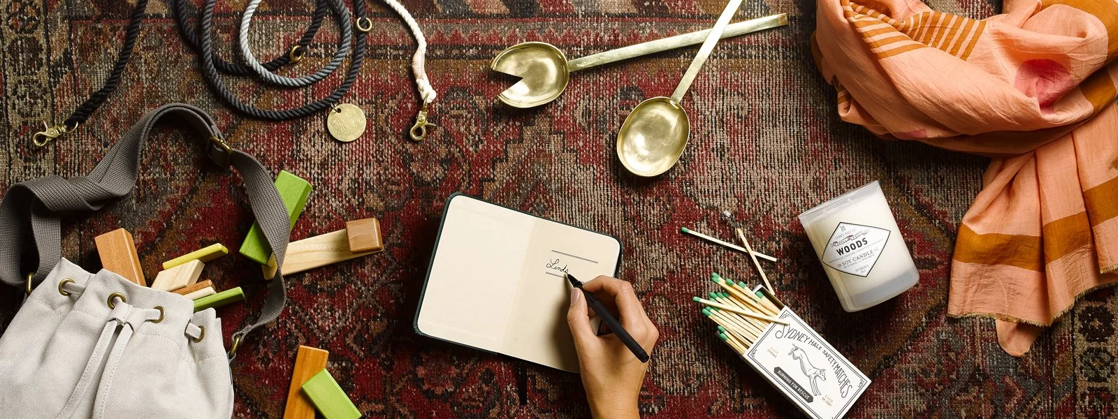 A collection of personal items on a patterned rug, including a beige tote bag, a notebook with a hand writing 'Lady' inside, a candle, a pair of gold spoons, various jewelry pieces, loosely folded pink and orange clothes, a box of matches, and a box