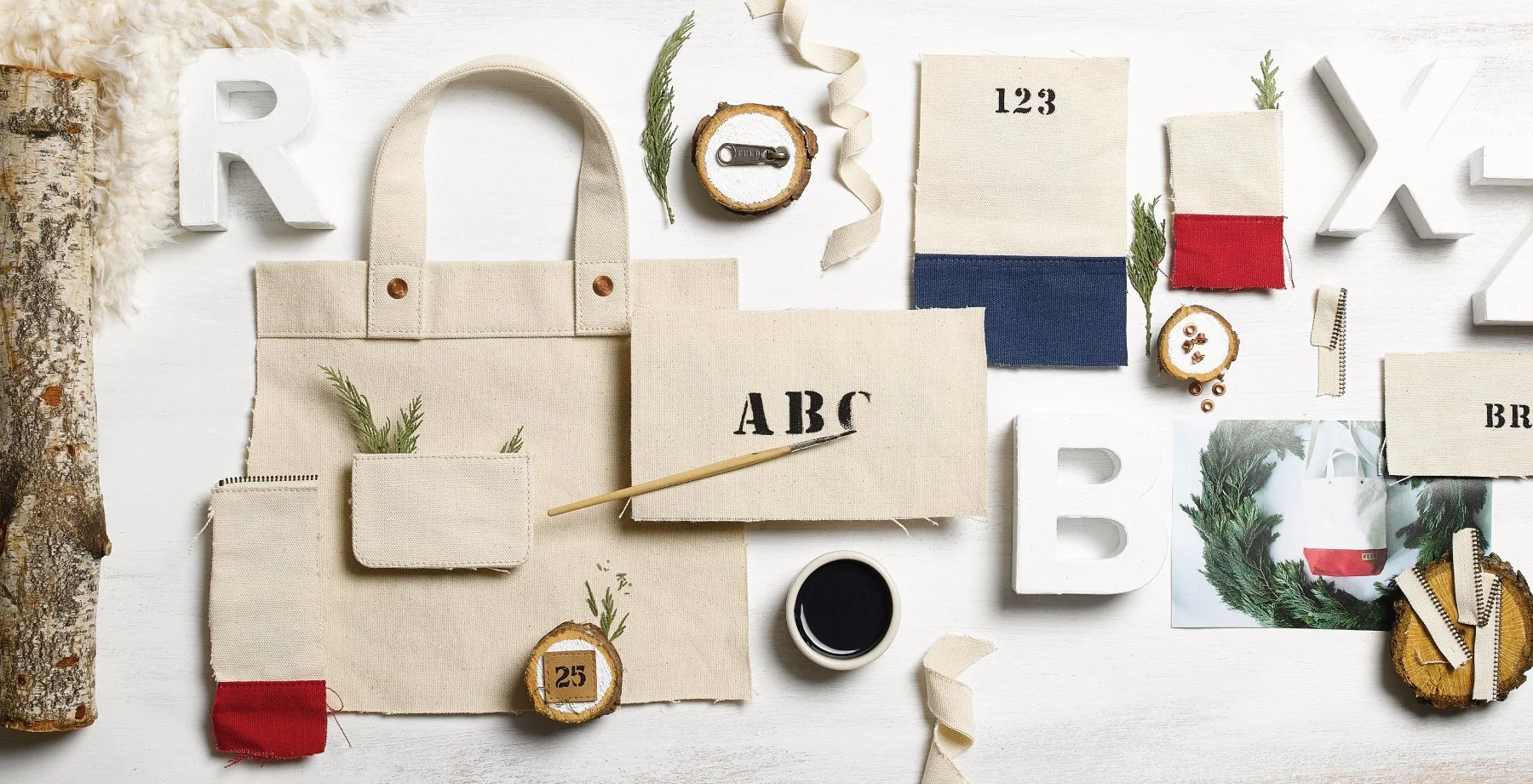 Crafting supplies for holiday decor on a white surface, including fabric in red, blue, and beige, wooden slices with numbers, greenery, zippers, a small cup of black paint, and decorative letters.