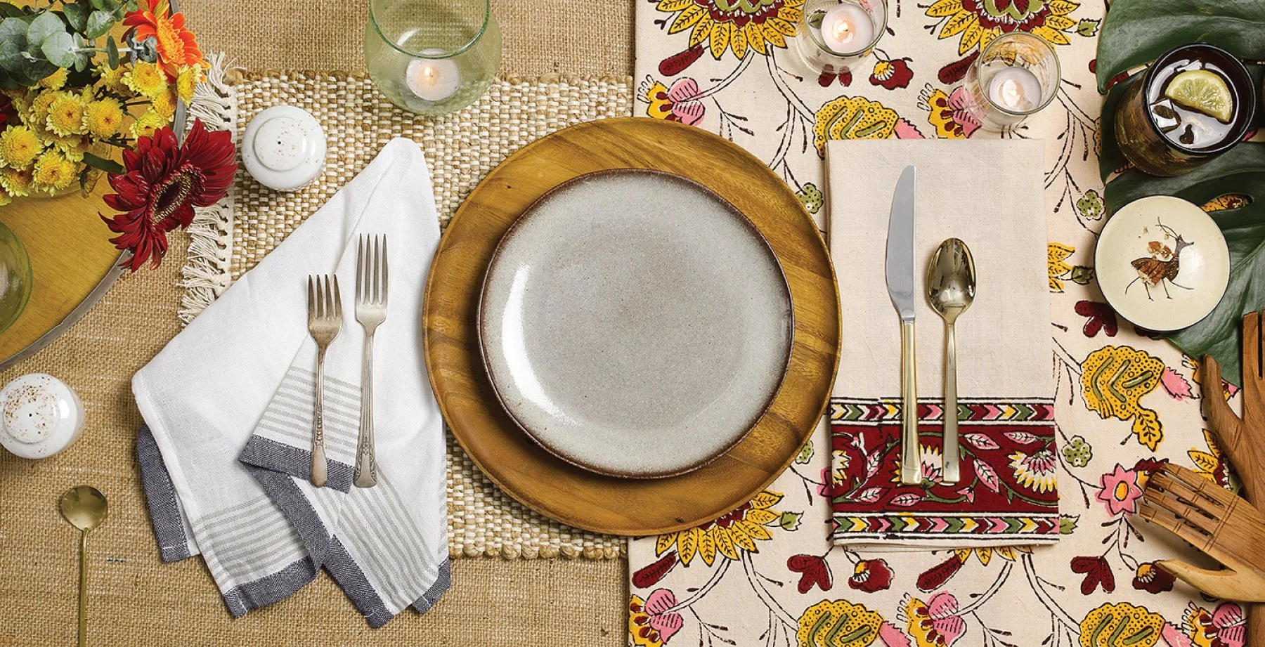 Table setting with a beige woven tablecloth, gold flatware, a gray and white cloth napkin, a round ceramic plate on a wooden charger, surrounded by glass candle holders, flowers, and seasonal decorations.