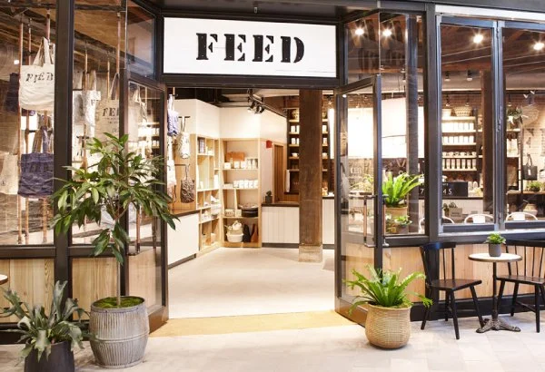 The storefront of a shop named FEED with glass doors and windows, potted plants outside, and a cozy interior visible inside with shelves and seating.