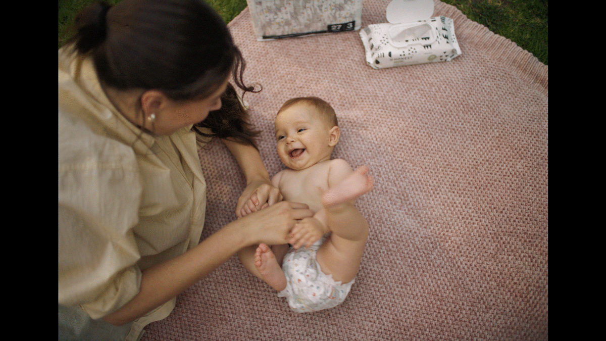 A woman and a baby are playing outside on a pink blanket, and the woman is holding the baby's foot while both are smiling and laughing.