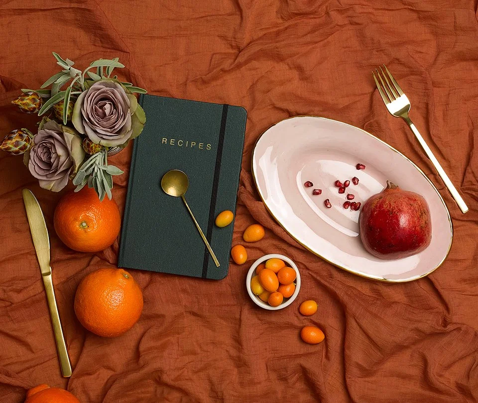 A table setting on a rust-colored cloth with a bouquet of purple roses and greenery, a dark green recipe book with a gold spoon on top, two orange tangerines, a small bowl of round orange fruits, an oval plate with a large pomegranate and pomegranate