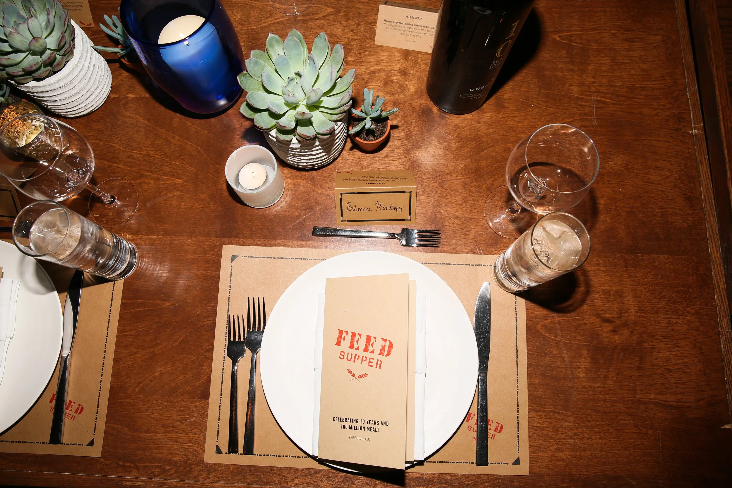 A dinner table set for a feeder supper event with a white plate, napkin, and stainless steel fork and knife, decorated with potted succulents, water glasses, a blue candleholder with a lit candle, and a small candle holder. There are brown placemats