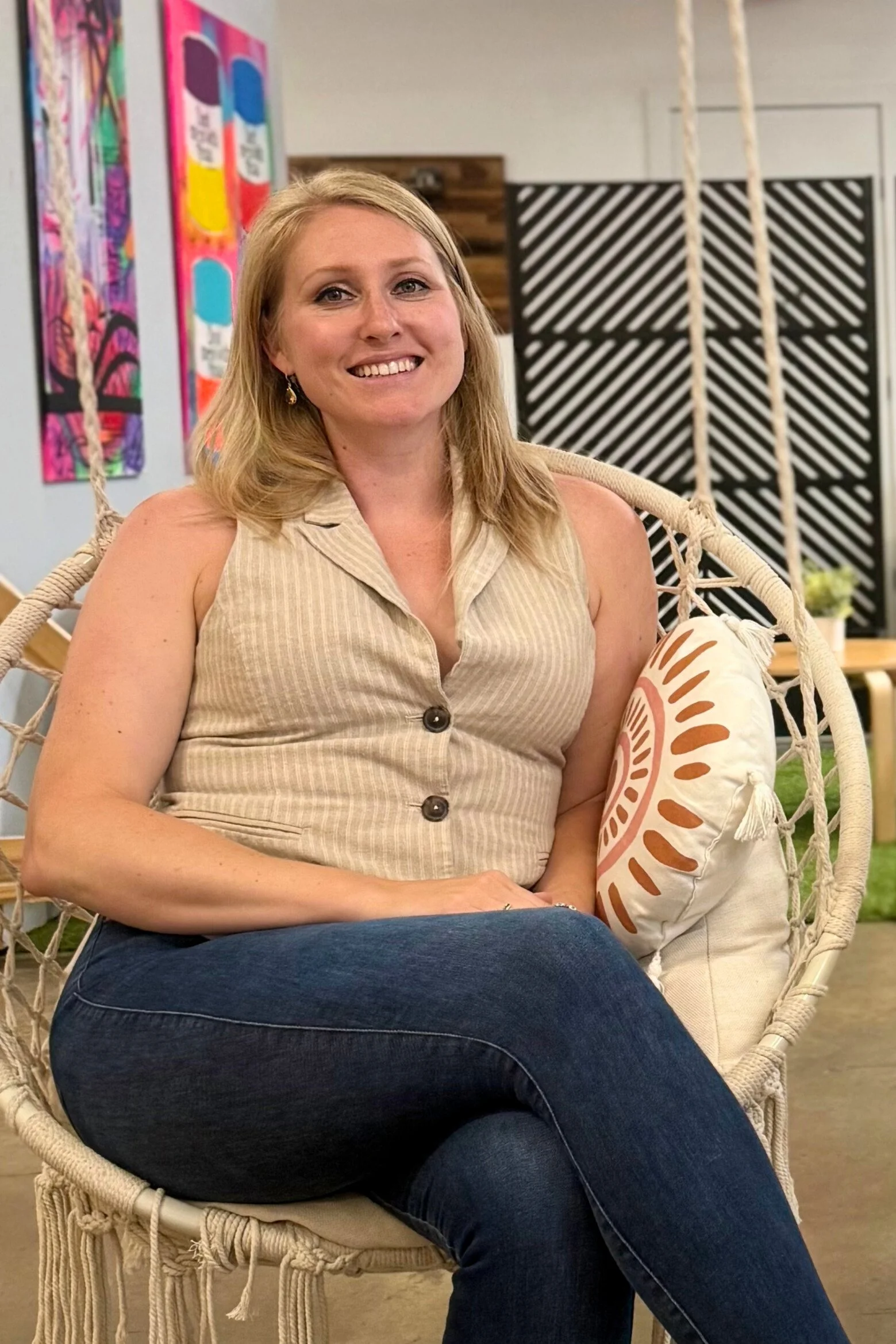 A smiling woman (co-founder Alexis Clark) with blonde hair sitting in a hanging basket chair with a decorative throw pillow, in a colorful, modern indoor space.